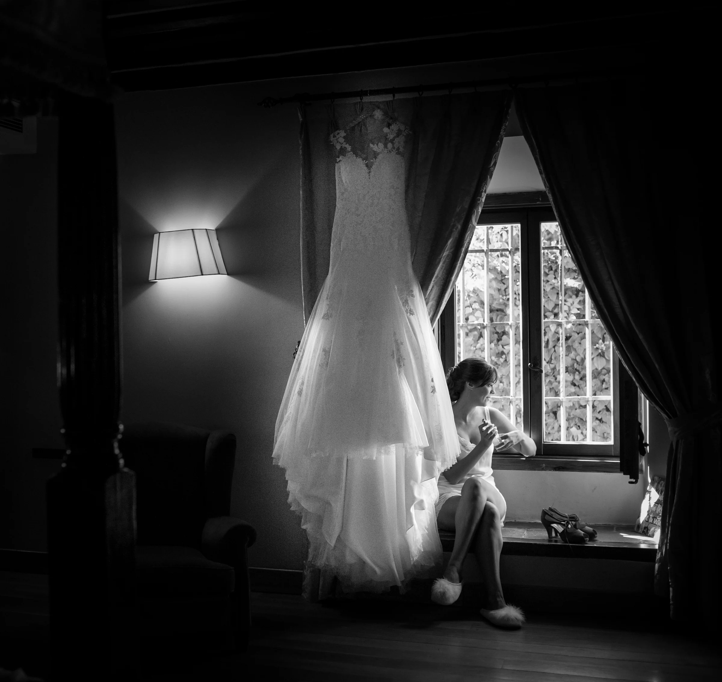 Vestido de novia colgado delante de la ventana mientras la novia se prepara en blanco y negro.