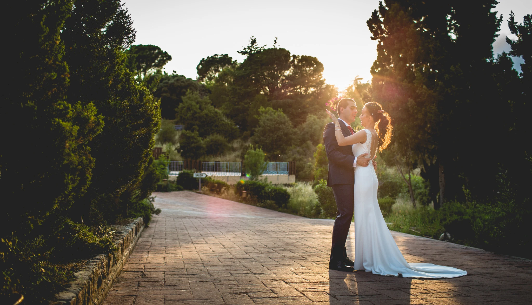 Novios abrazados en un camino con el sol del atardecer iluminando la escena.