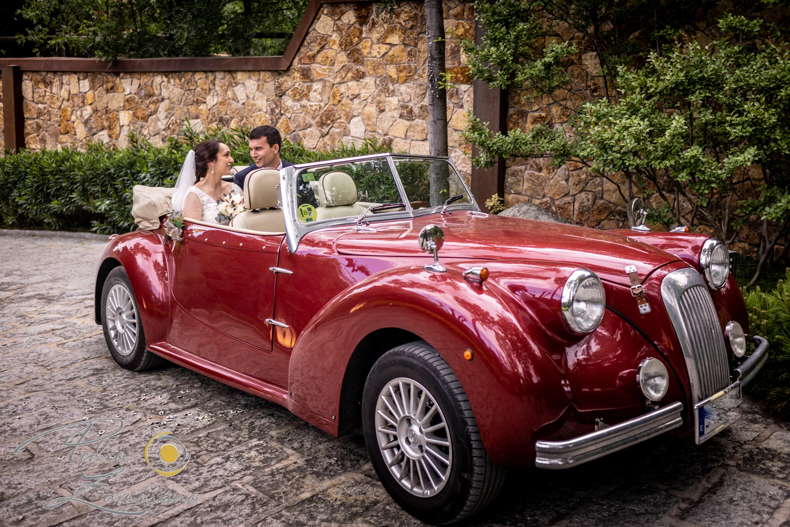 Pareja de novios junto a un coche clásico rojo en los jardines de Hacienda Jacaranda.