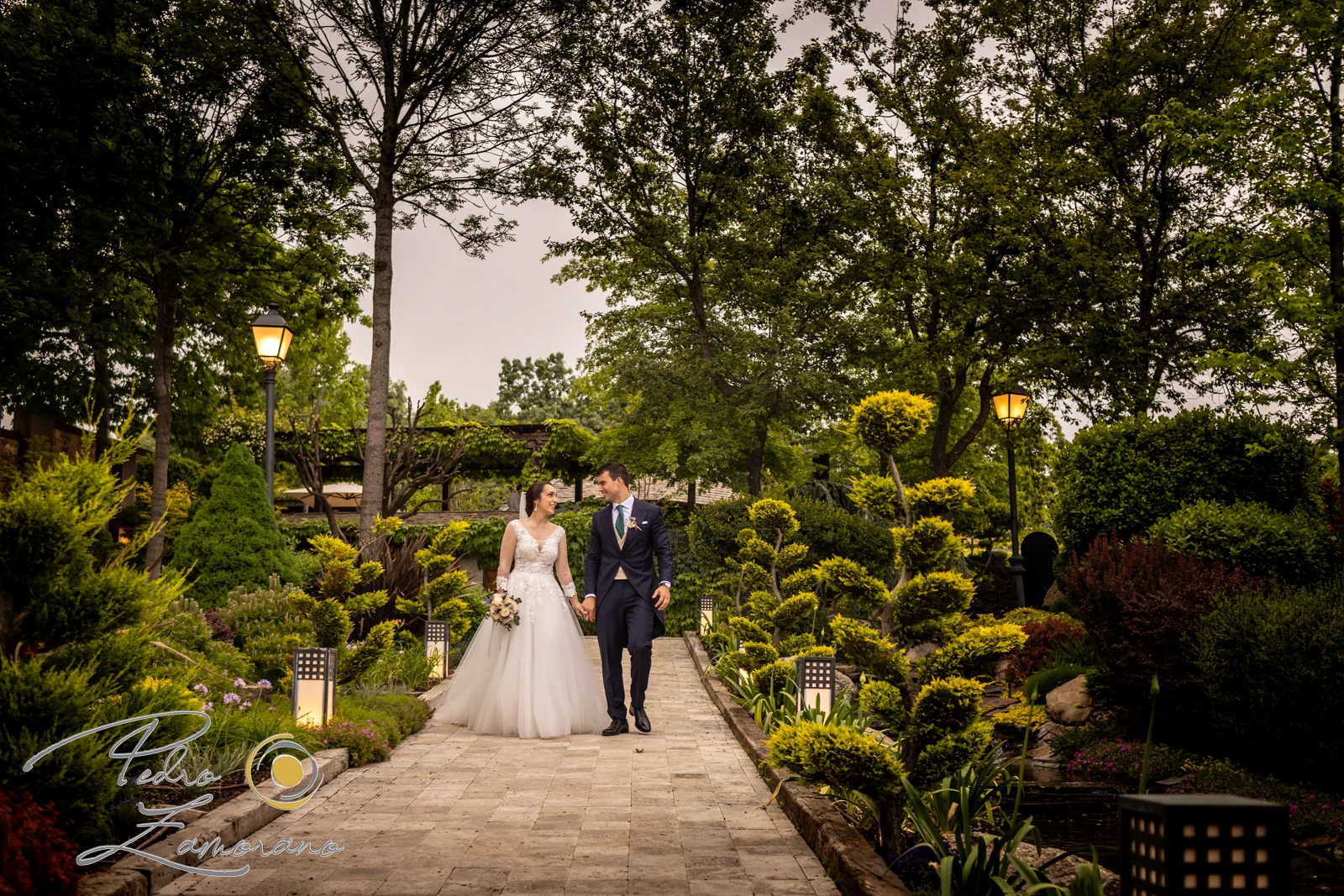 Novios paseando por un camino ajardinado junto al estanque de Hacienda Jacaranda.