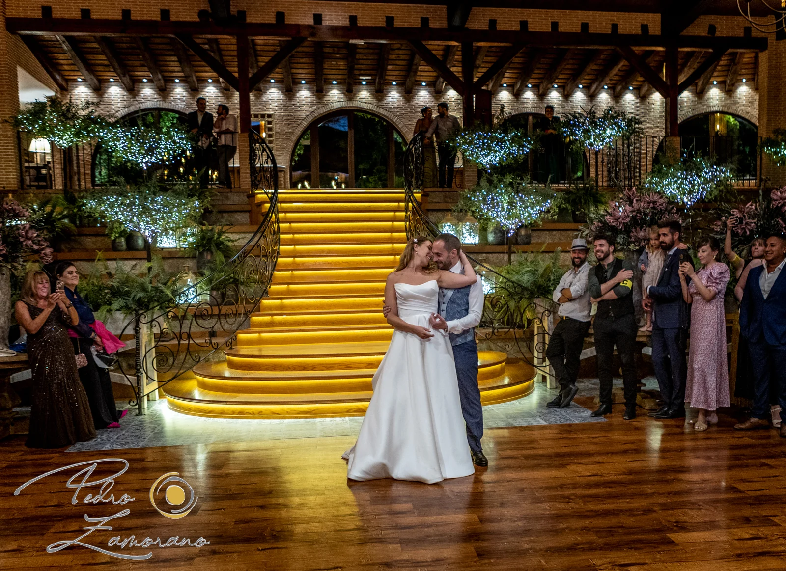 Novios bailando con la escalera iluminada del salón de Hacienda Jacaranda al fondo.