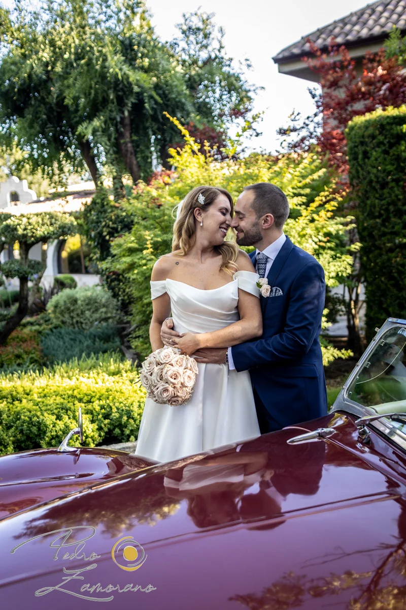 Pareja de recién casados junto a un coche clásico rojo en Hacienda Jacaranda.