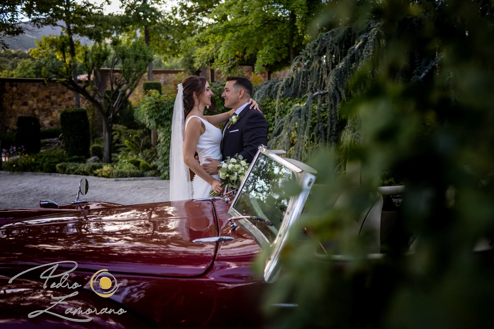 Novios junto a un coche clásico rojo fotografiados a través de la vegetación de Hacienda Jacaranda.