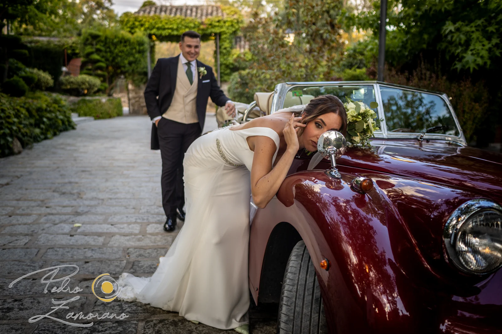 Novia apoyada en un coche clásico rojo mientras el novio se acerca en Hacienda Jacaranda.