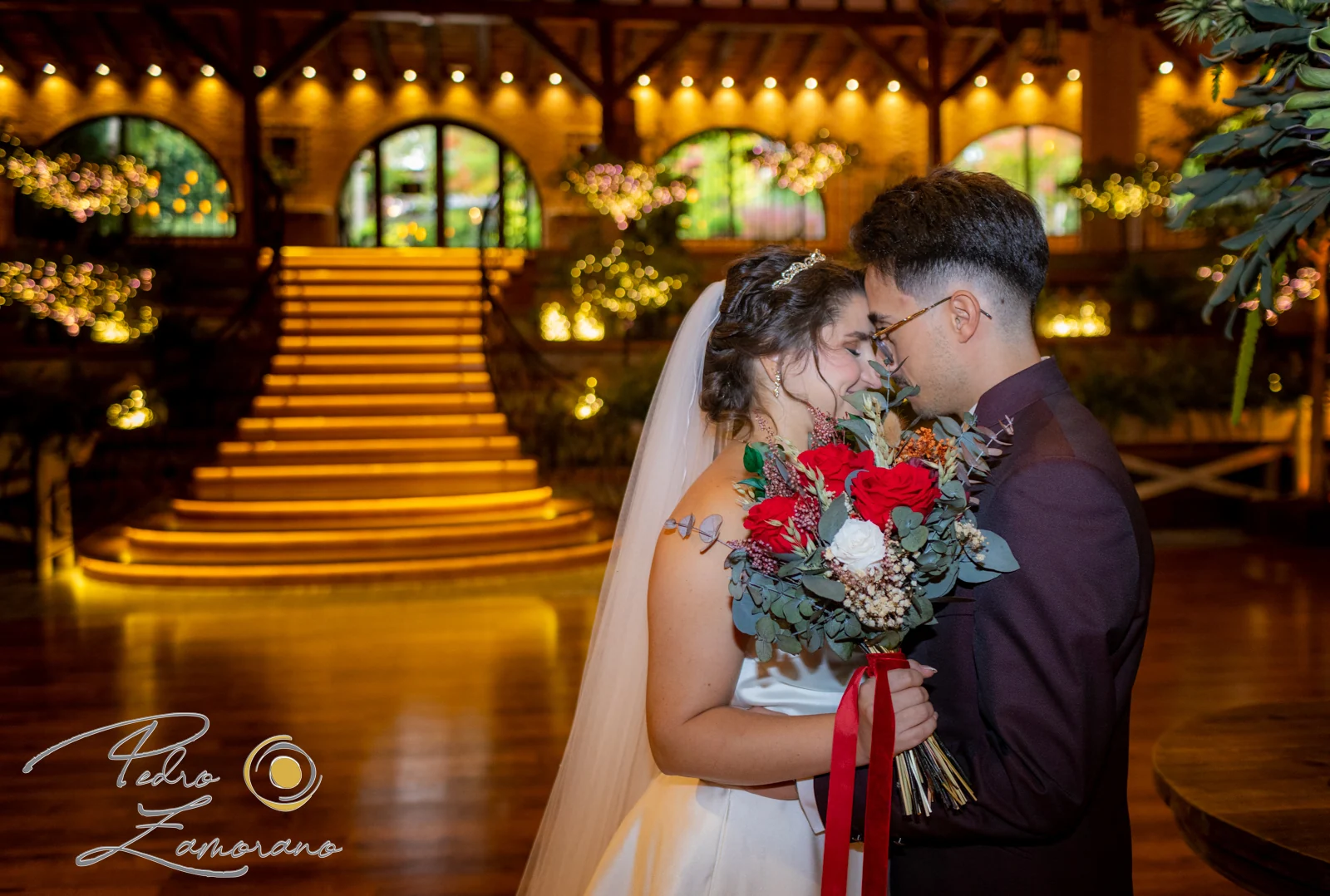 Pareja abrazada con un ramo de flores rojas y la escalera iluminada de Hacienda Jacaranda al fondo.