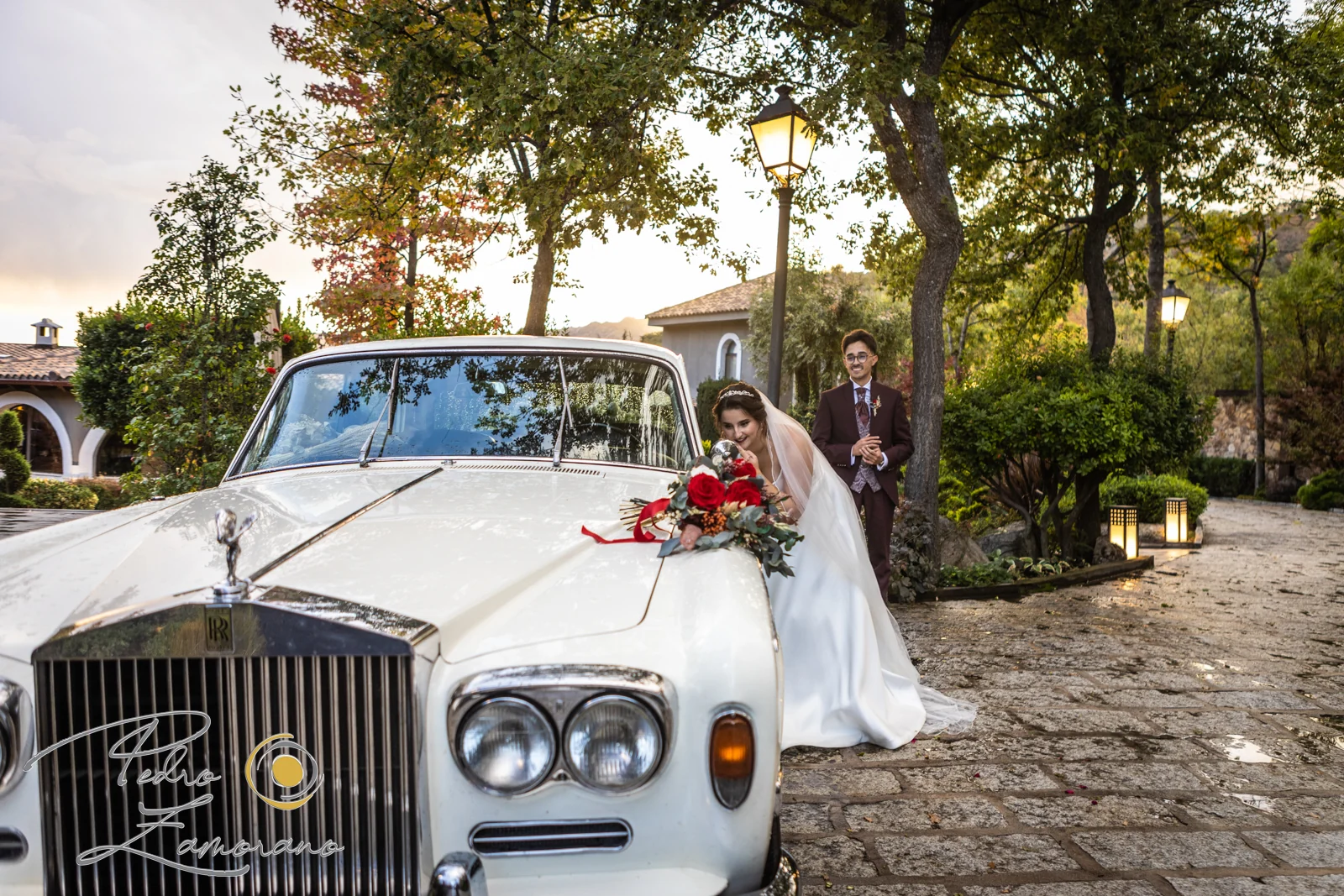 Novia junto a un Rolls-Royce blanco clásico con el novio al fondo en los jardines de Hacienda Jacaranda.