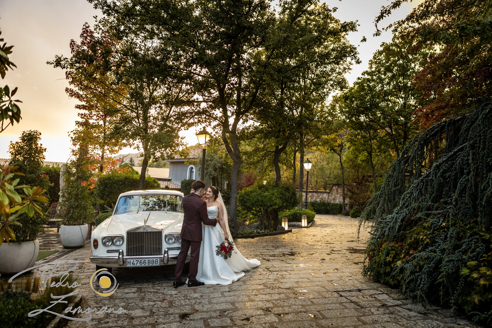 Novios junto a un Rolls-Royce blanco al atardecer en los jardines de Hacienda Jacaranda.
