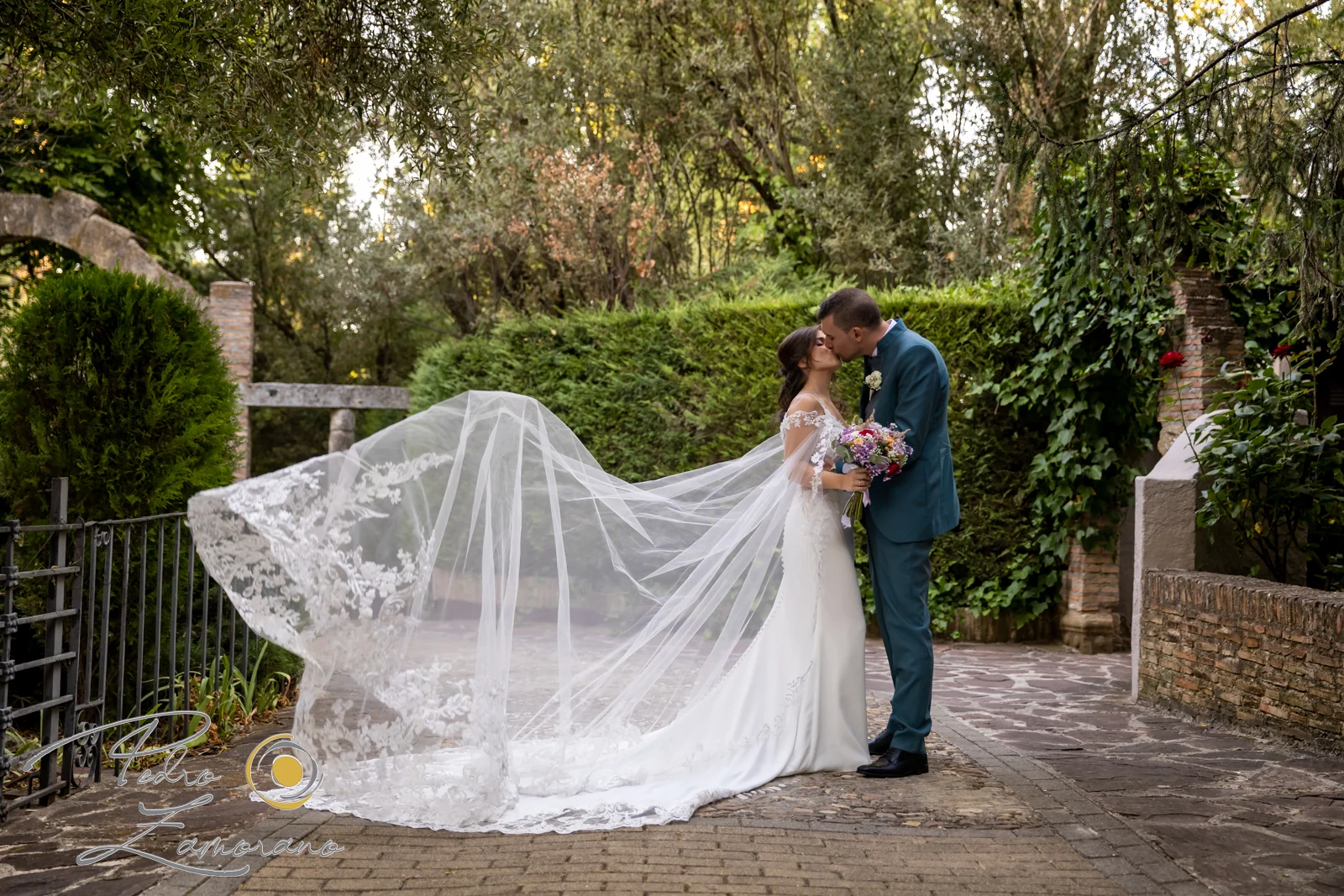 Novios besándose en los jardines de Jardín El Botero con el velo de la novia extendido al viento.
