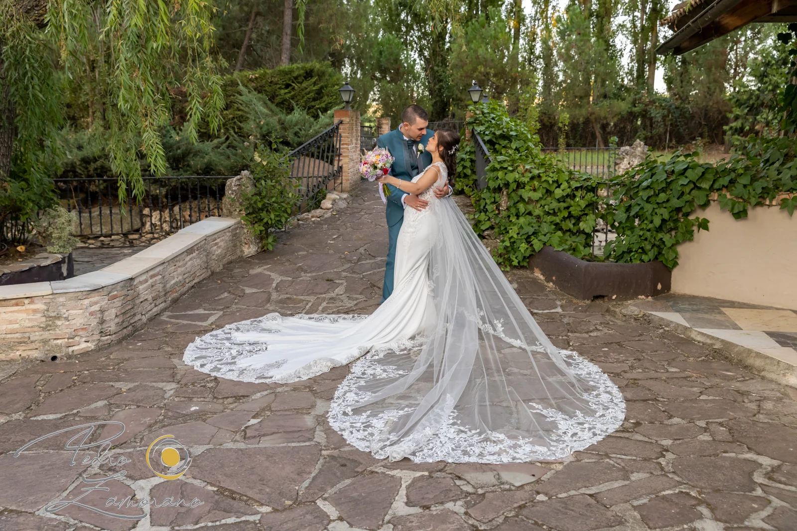 Novios abrazados en un camino de piedra con el largo velo de la novia extendido por los jardines de Jardín El Botero.