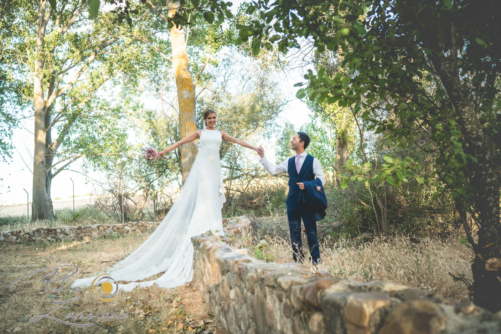 Novios cogidos de la mano junto a un muro de piedra entre árboles en Jardín El Botero.