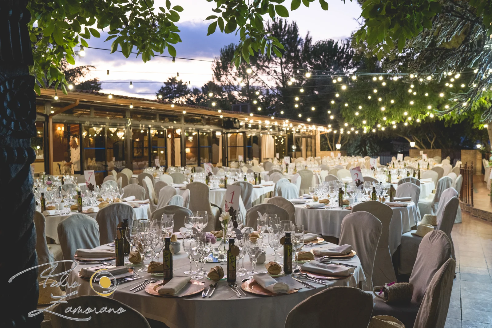Mesas preparadas para una boda bajo guirnaldas de luces al anochecer en Jardín El Botero.
