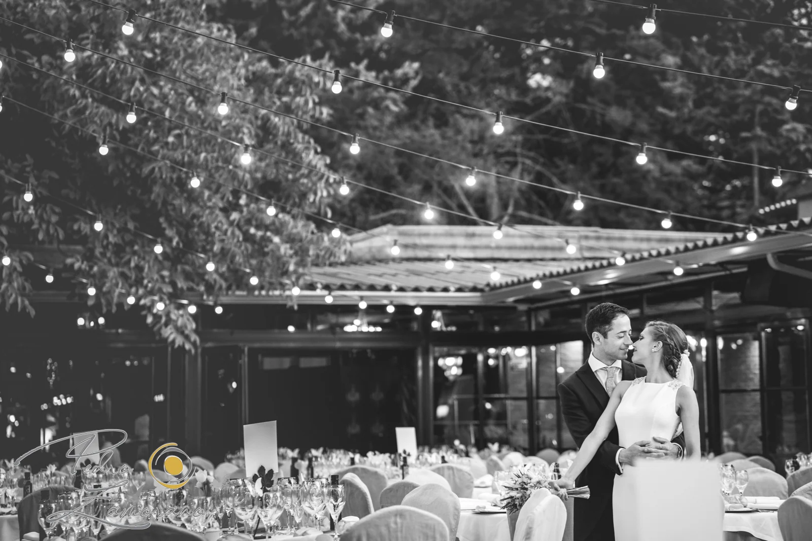 Novios abrazados entre las mesas y las guirnaldas de luces de Jardín El Botero en una fotografía en blanco y negro.