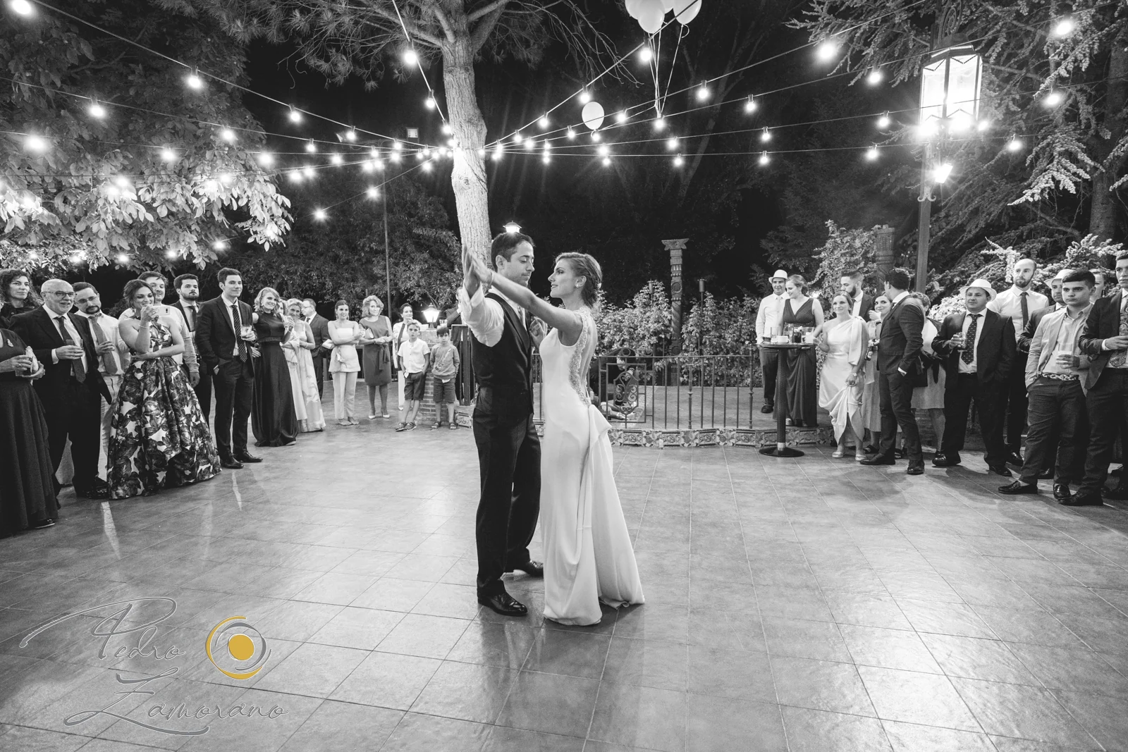 Novios bailando rodeados por sus invitados bajo las luces de Jardín El Botero en blanco y negro.