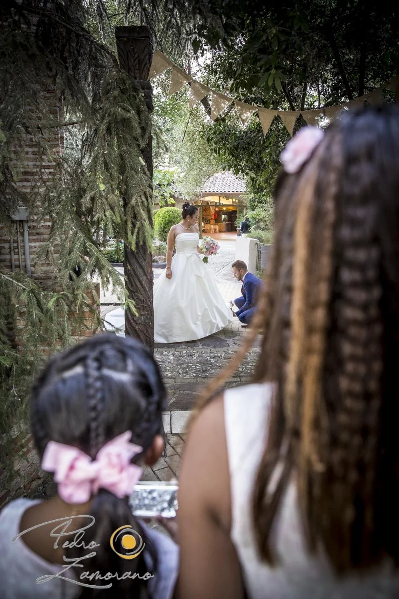 Novia en un camino ajardinado mientras el novio se agacha para ayudarle con el vestido en Jardín El Botero.