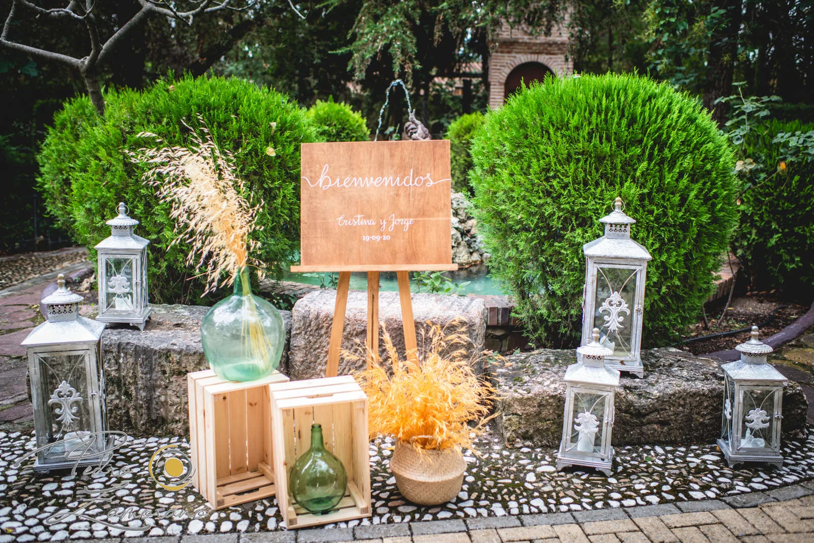 Decoración de bienvenida con cartel de boda y faroles junto a una fuente en los jardines de Jardín El Botero.