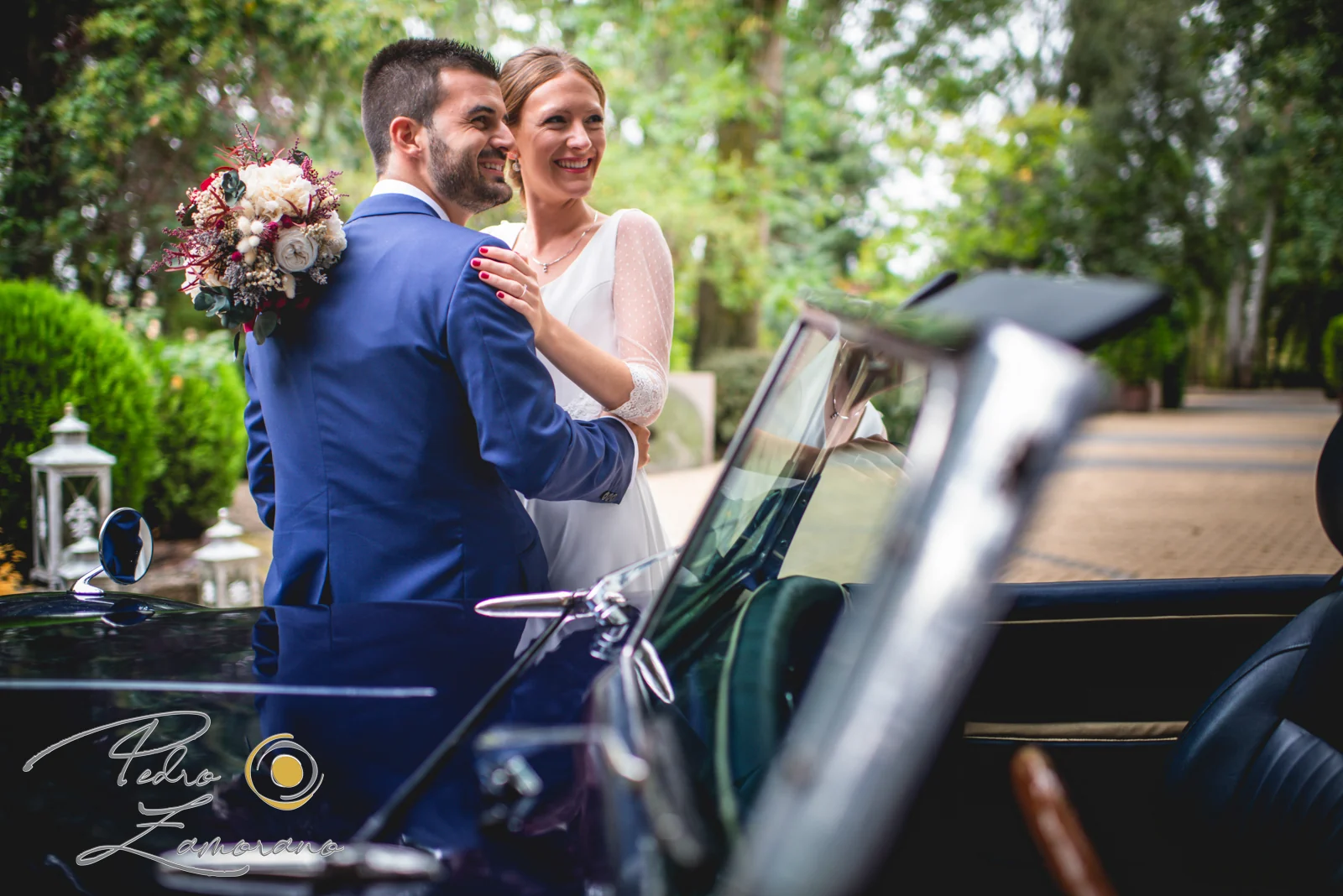 Novios abrazados junto a un coche clásico descapotable en los jardines de Jardín El Botero.