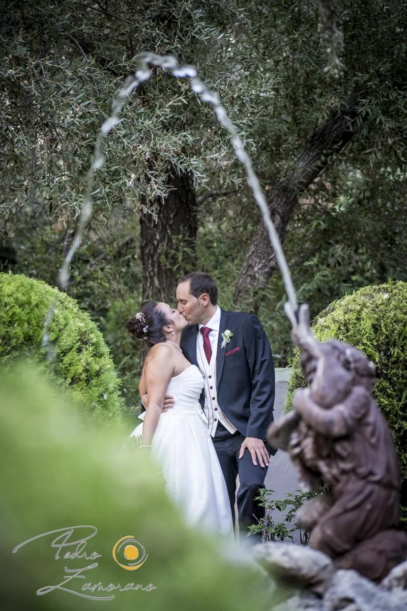 Novios besándose en los jardines de Jardín El Botero enmarcados por el arco de agua de una fuente.