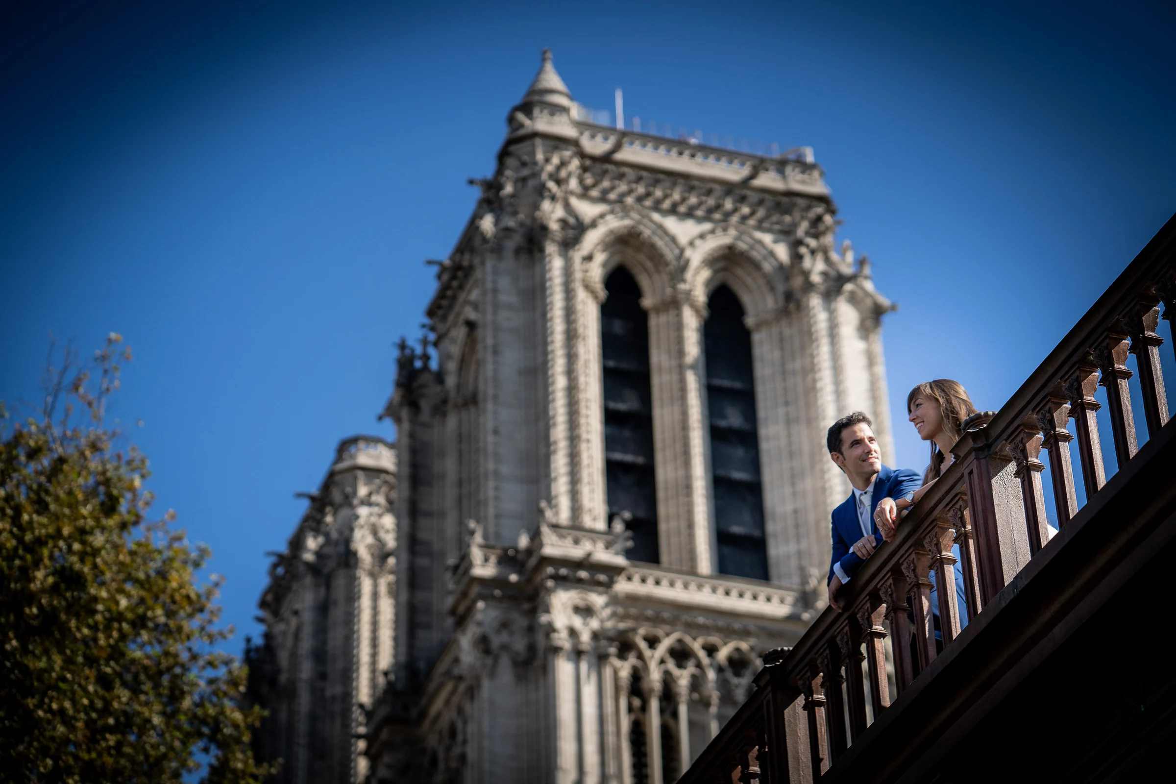 José Luis y Lara conversan sobre un puente con una de las torres de Notre-Dame elevándose detrás.