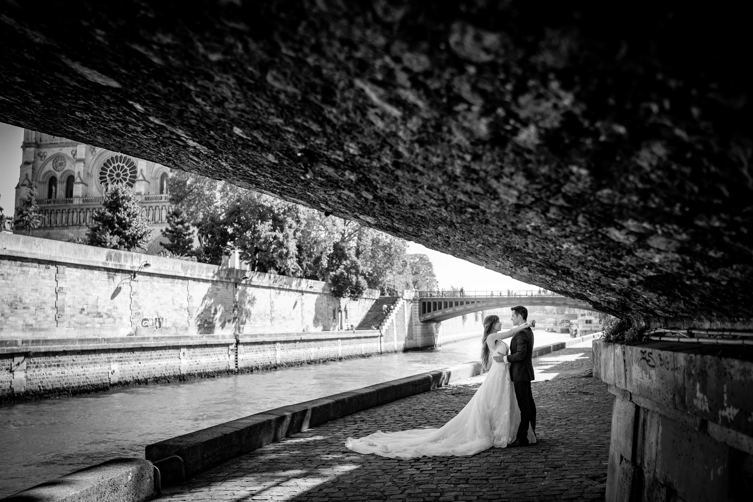 José Luis y Lara abrazados bajo un puente junto al Sena en una fotografía panorámica en blanco y negro.