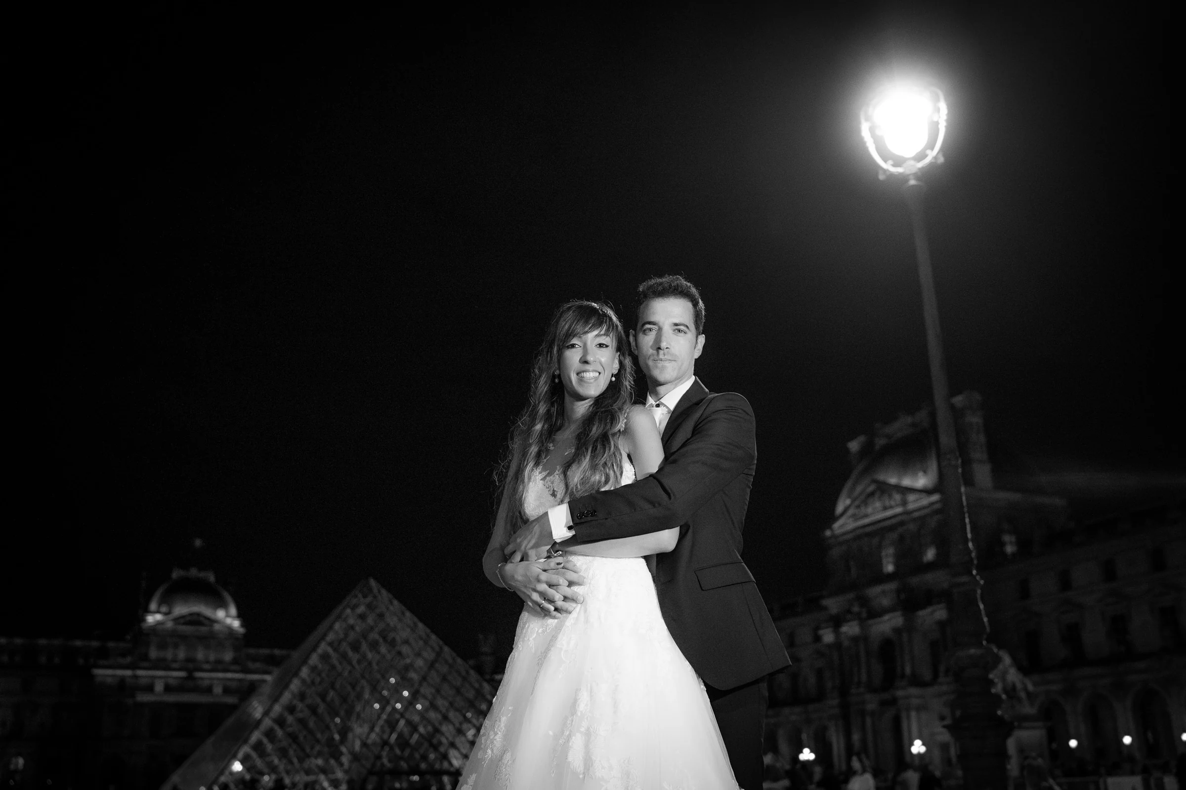 José Luis y Lara frente al Louvre de noche en una fotografía en blanco y negro con una farola iluminada.