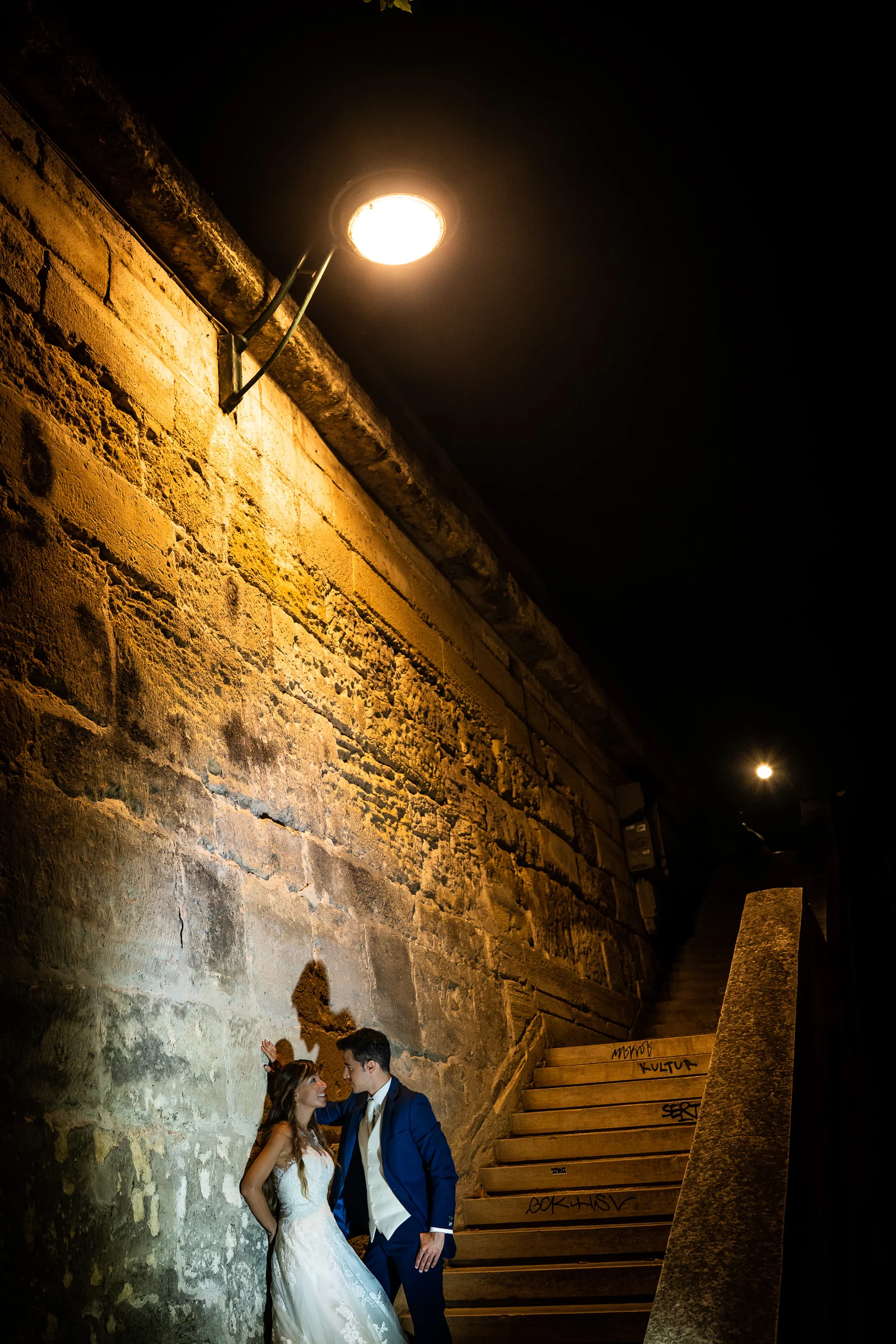José Luis y Lara junto a unas escaleras y un muro iluminado durante la noche de su postboda en París.
