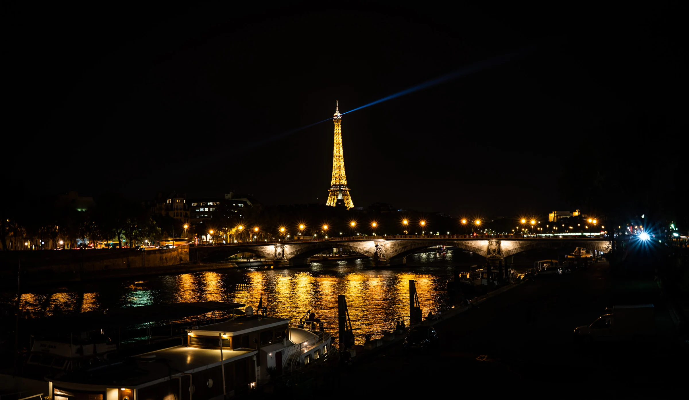 Panorámica nocturna del Sena con un puente iluminado y la Torre Eiffel proyectando su haz de luz sobre París.