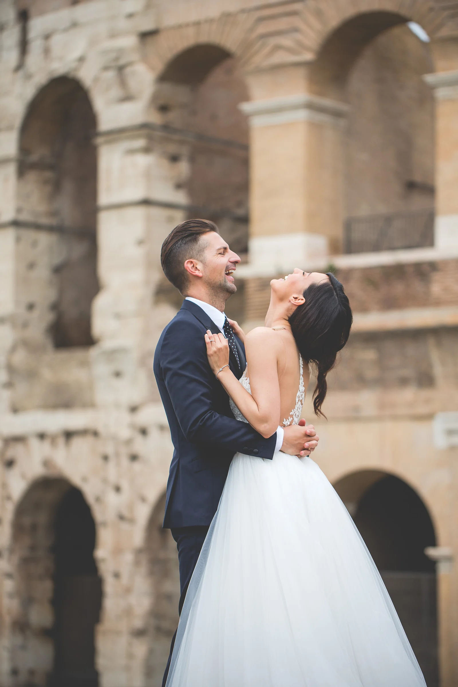 El novio abraza y besa a la novia con los arcos del Coliseo desenfocados al fondo durante una sesión postboda en Coliseo de Roma.