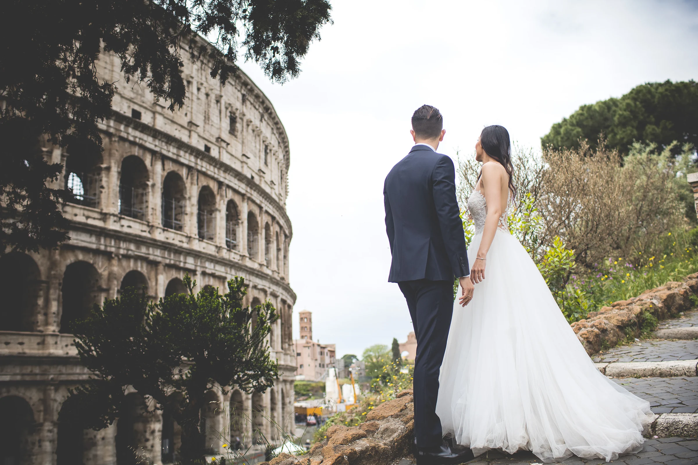 Los novios caminan de espaldas junto al Coliseo y la vegetación del entorno durante una sesión postboda en Coliseo de Roma.