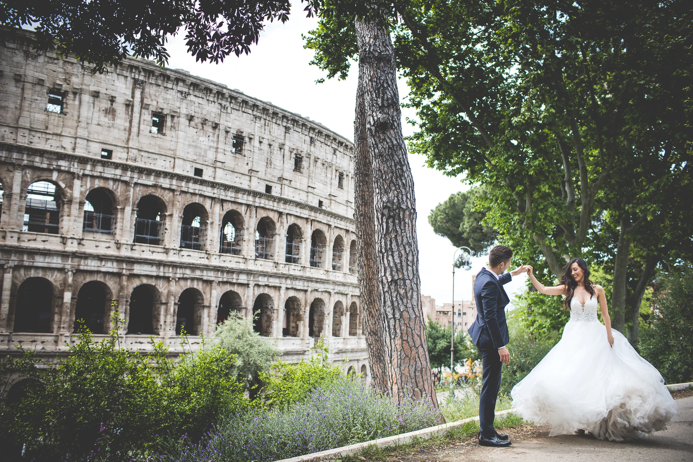 La pareja conversa junto a un árbol con el Coliseo enmarcando la escena durante una sesión postboda en Coliseo de Roma.