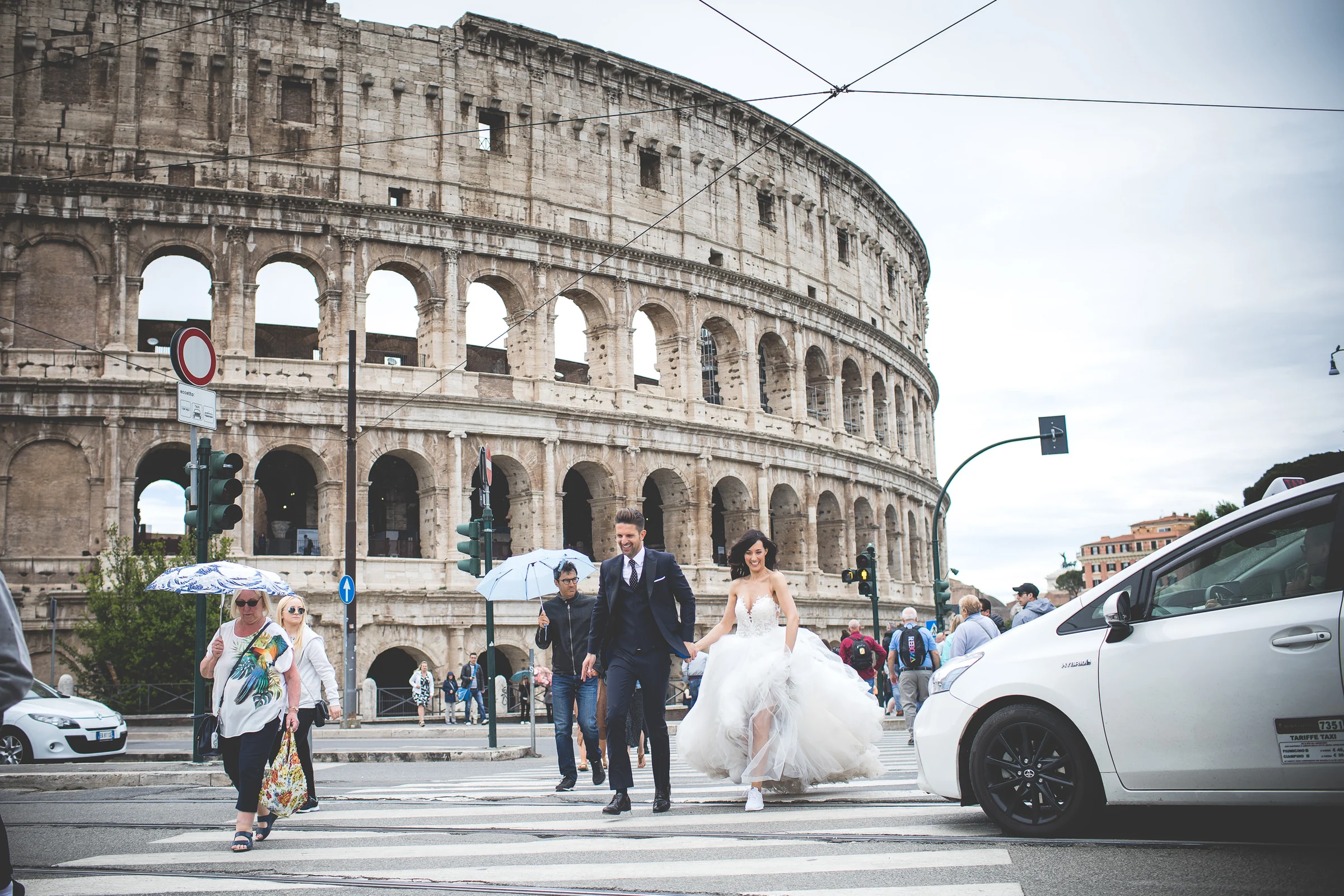 Los novios cruzan una calle concurrida junto al Coliseo durante la sesión durante una sesión postboda en Coliseo de Roma.