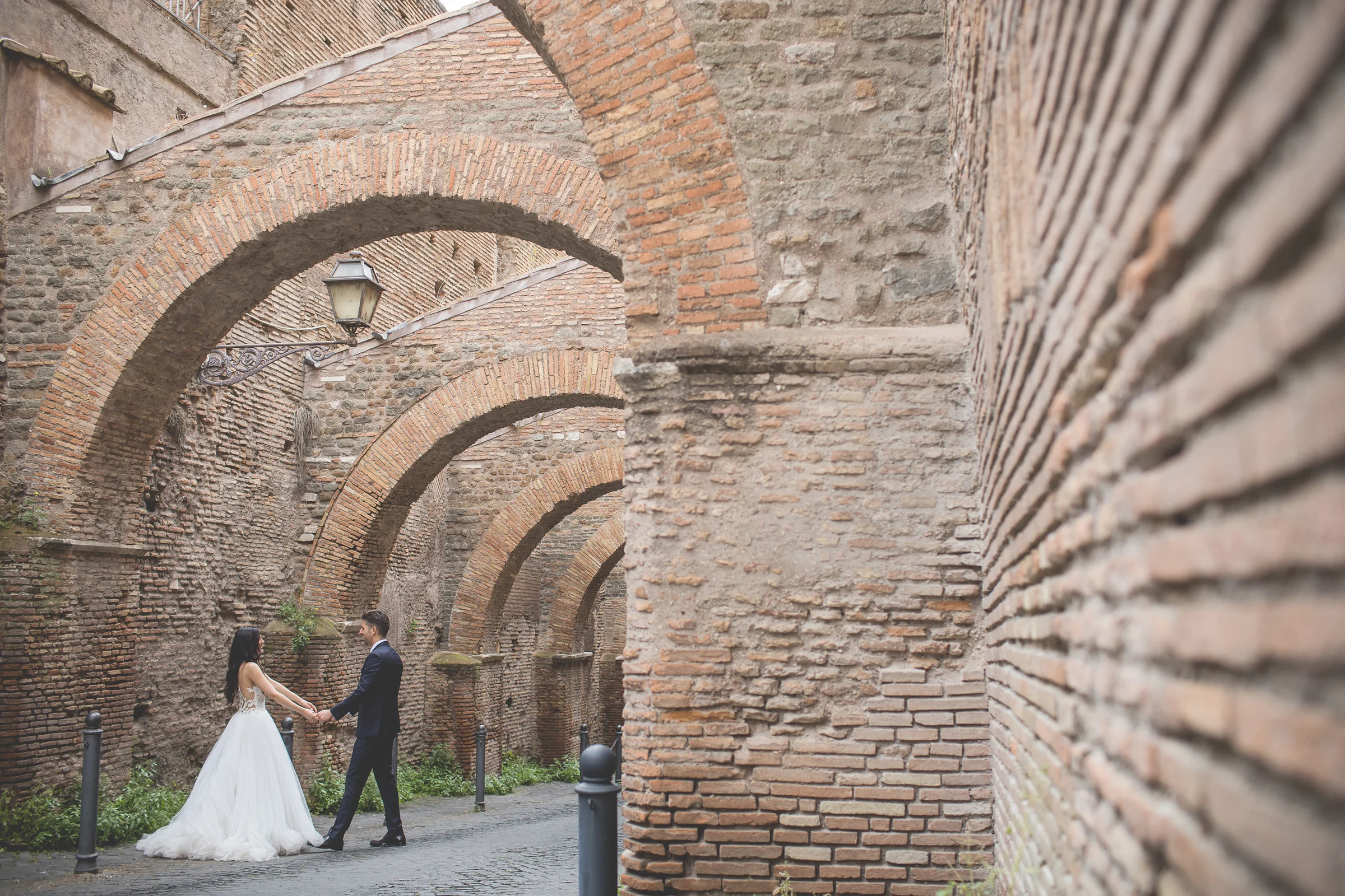 Los novios caminan bajo una sucesión de arcos de ladrillo romanos durante una sesión postboda en Foro Romano y área arqueológica.