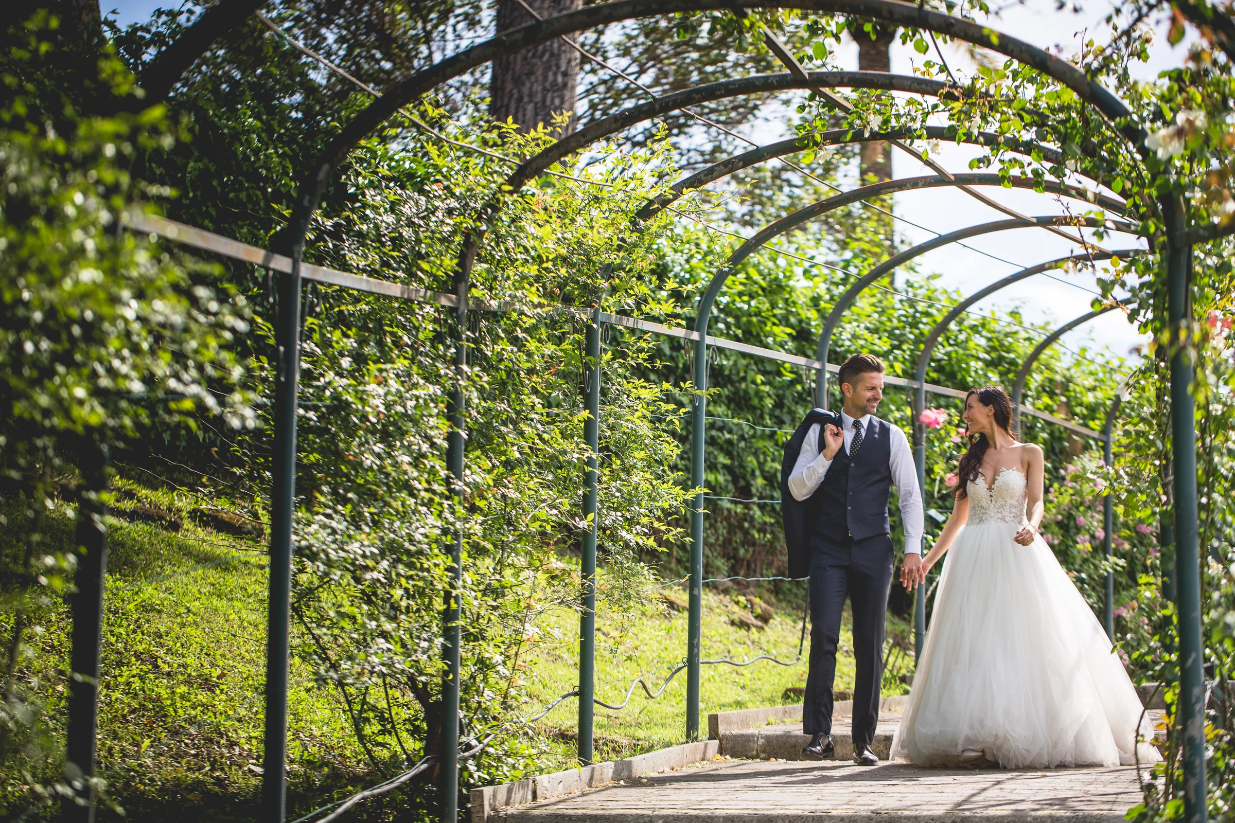 La pareja camina bajo una pérgola verde en los jardines de Roma durante una sesión postboda en Jardines y miradores de Roma.
