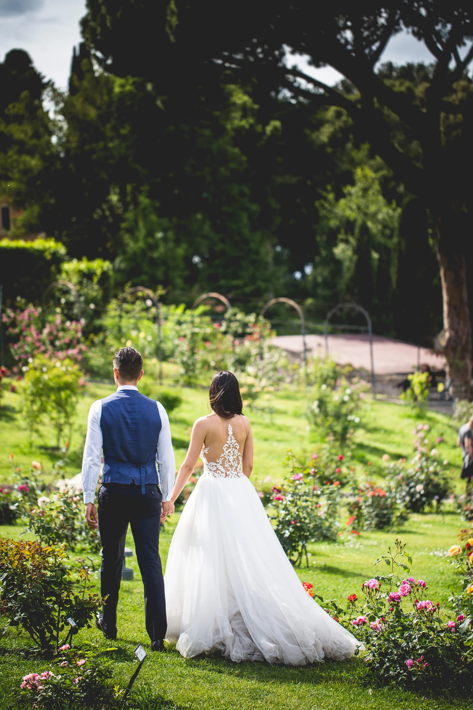 La pareja camina de espaldas por un jardín arbolado de Roma durante una sesión postboda en Jardines y miradores de Roma.