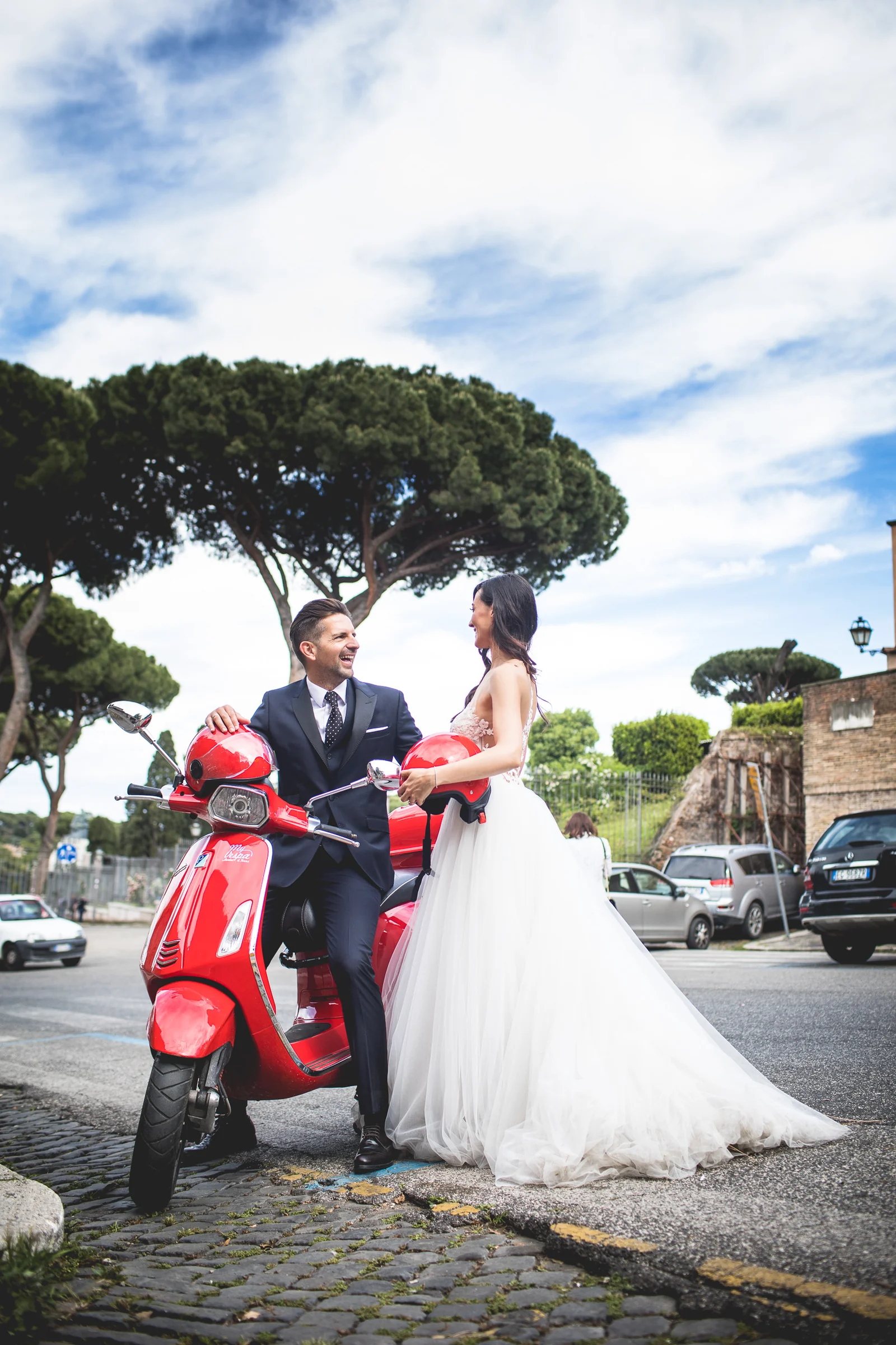 Los novios posan junto a una Vespa roja bajo pinos de Roma durante una sesión postboda en Calles de Roma con Vespa.