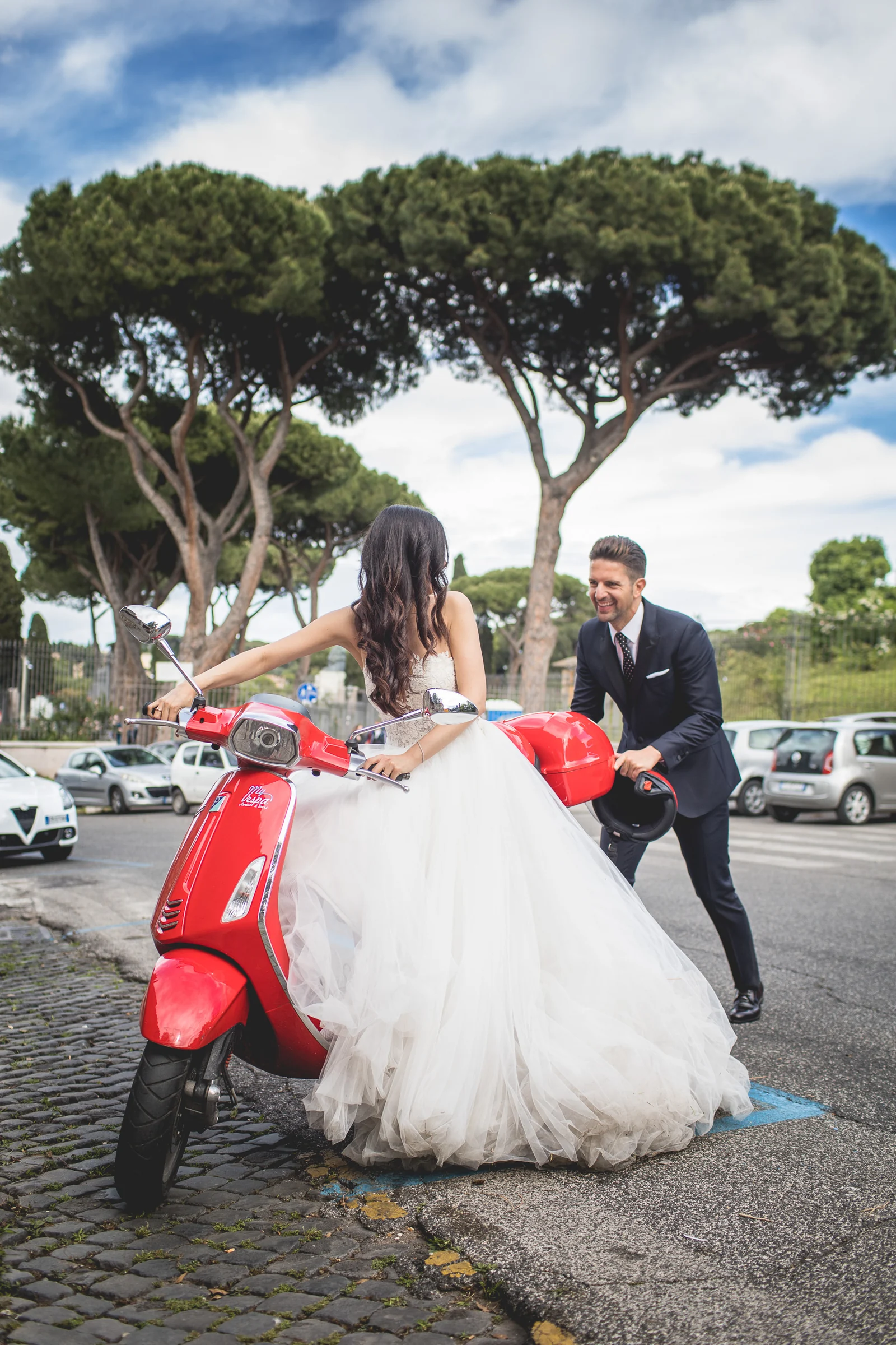 Los novios posan con una Vespa roja en una calle arbolada de Roma durante una sesión postboda en Calles de Roma con Vespa.