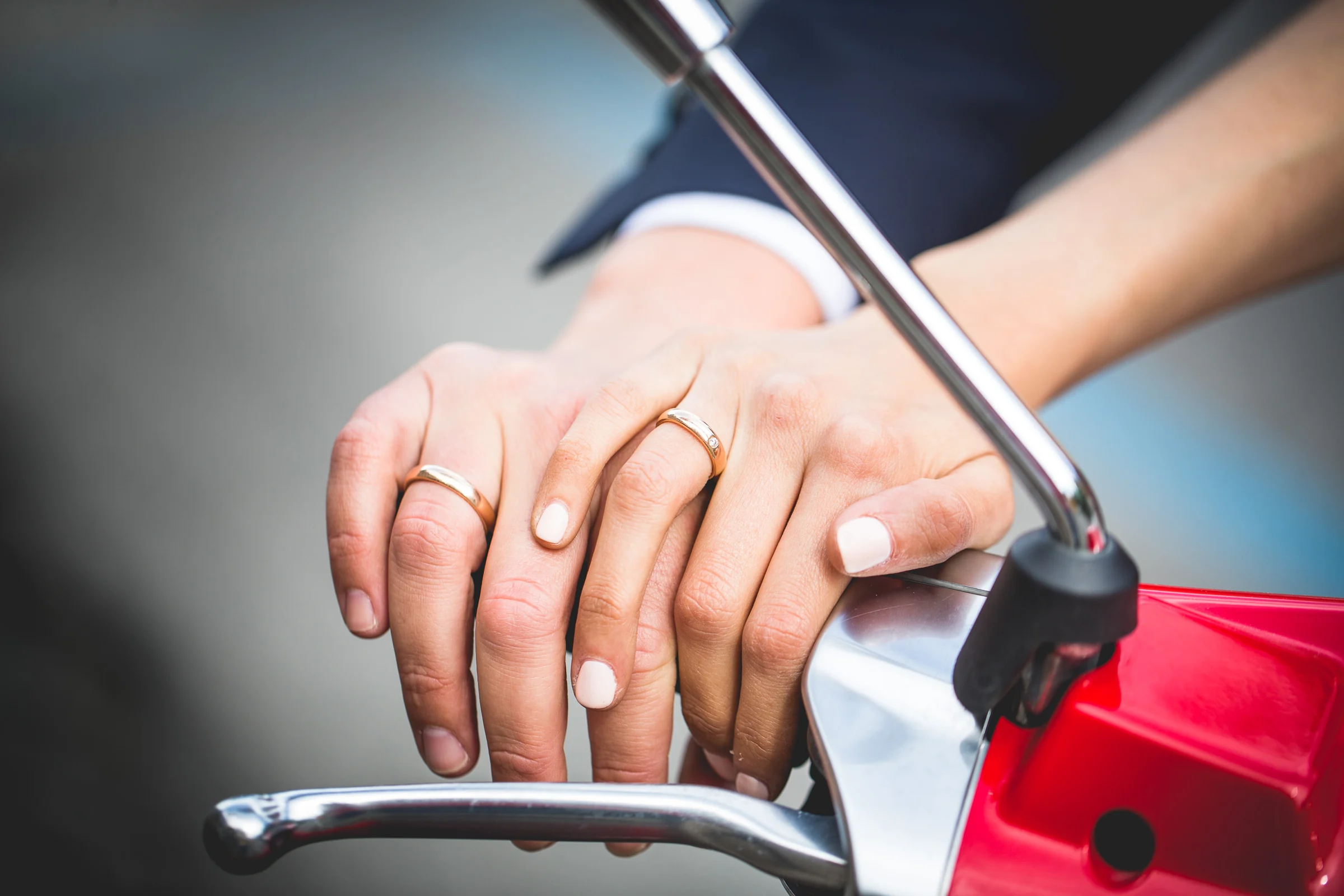Detalle de las manos de la pareja unidas sobre el manillar de una Vespa roja durante una sesión postboda en Calles de Roma con Vespa.
