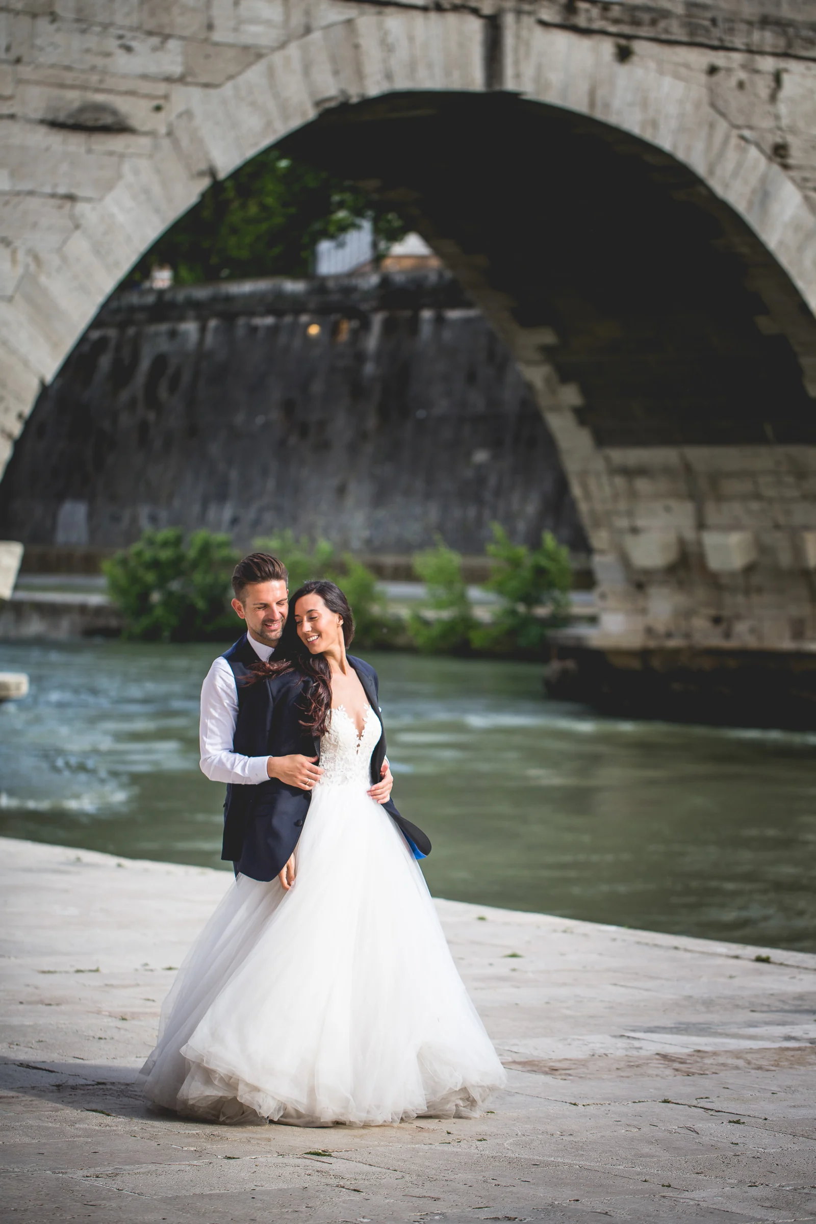 Los novios se abrazan junto al río bajo un gran arco de piedra durante una sesión postboda en Río Tíber y puentes de Roma.