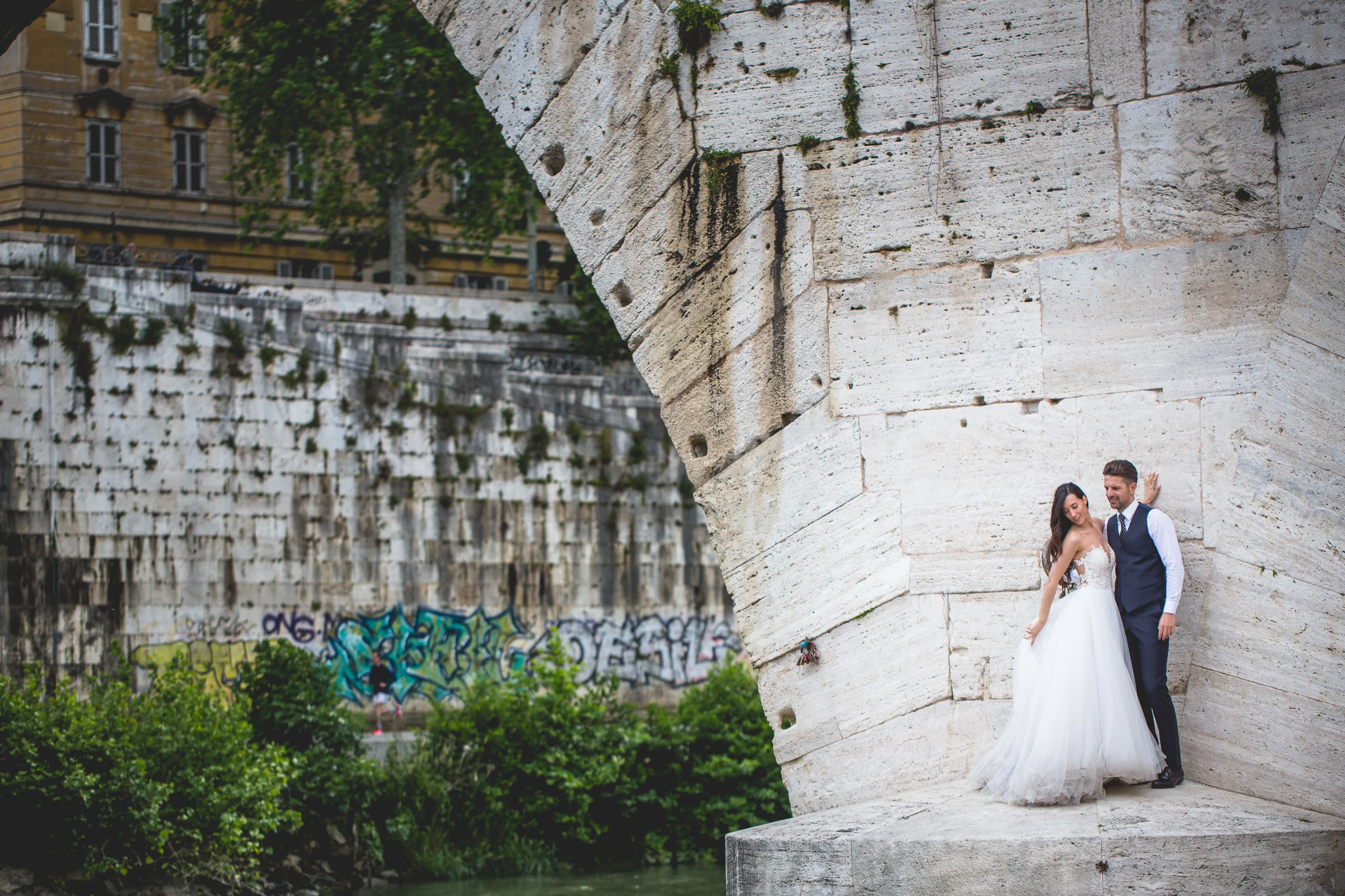 Los novios posan bajo un arco de puente con grafitis y vegetación durante una sesión postboda en Río Tíber y puentes de Roma.