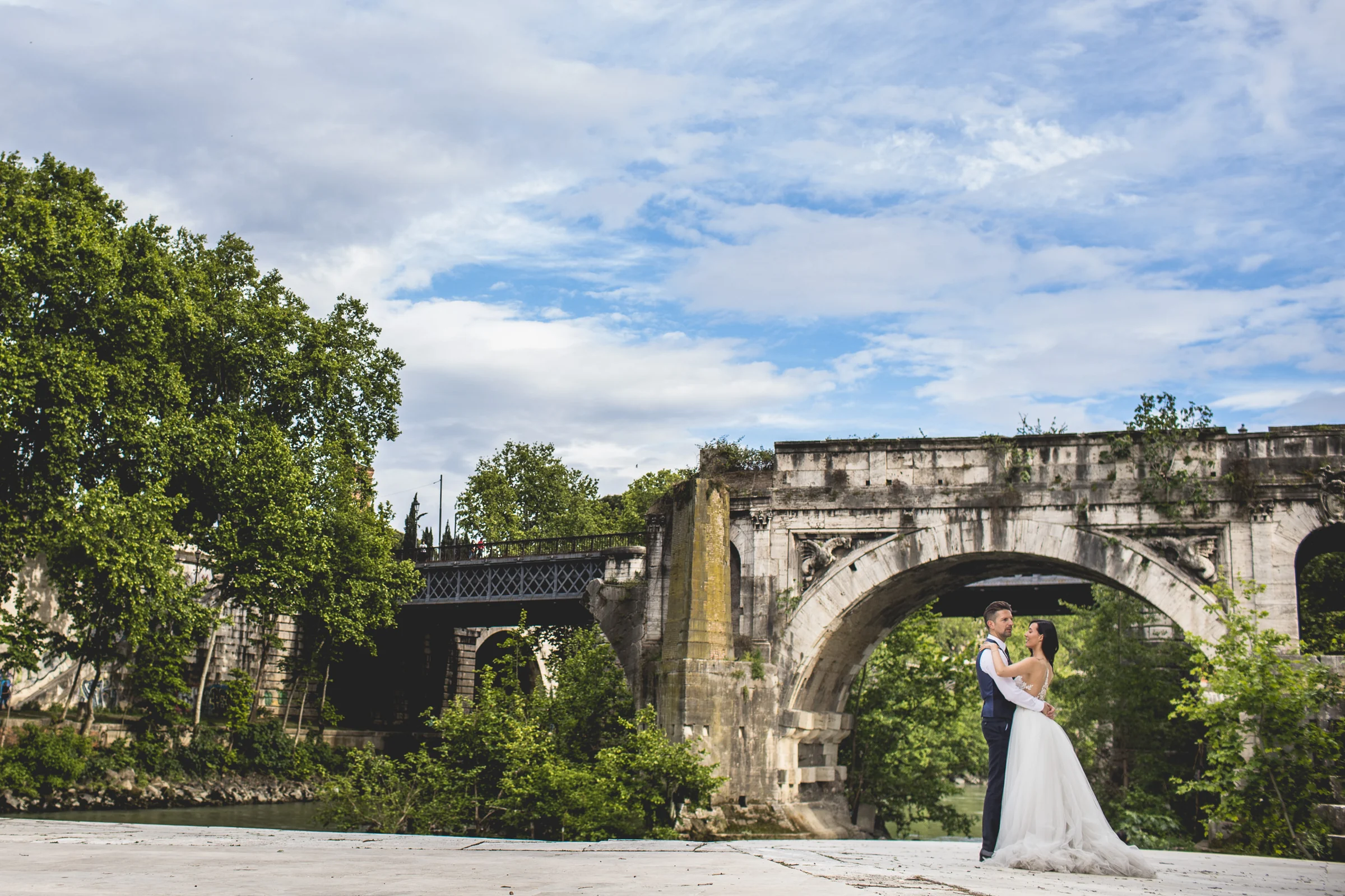 Vista panorámica de la pareja junto al Tíber y un puente histórico de Roma durante una sesión postboda en Río Tíber y puentes de Roma.