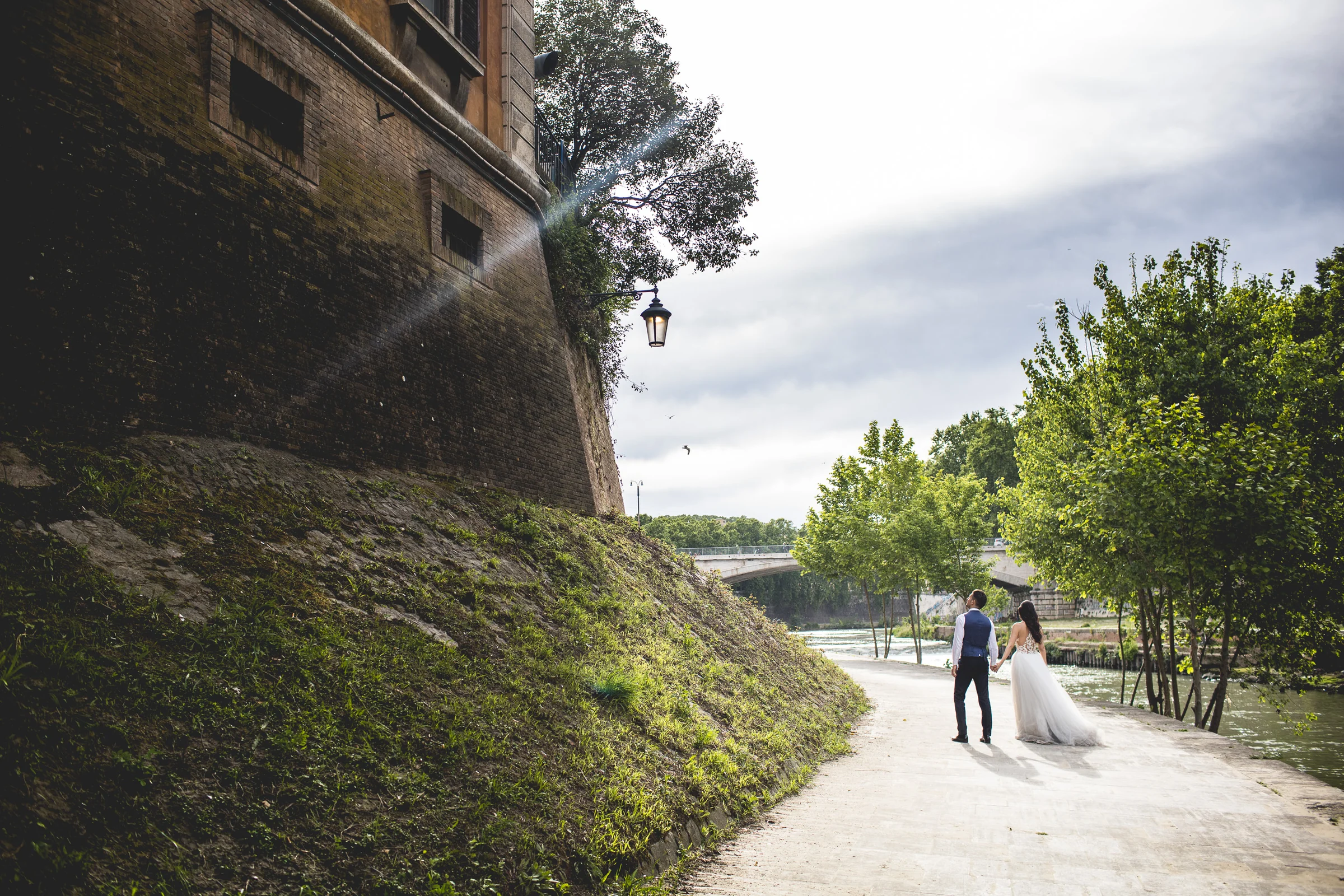 Los novios caminan por un paseo arbolado a orillas del río durante una sesión postboda en Río Tíber y puentes de Roma.