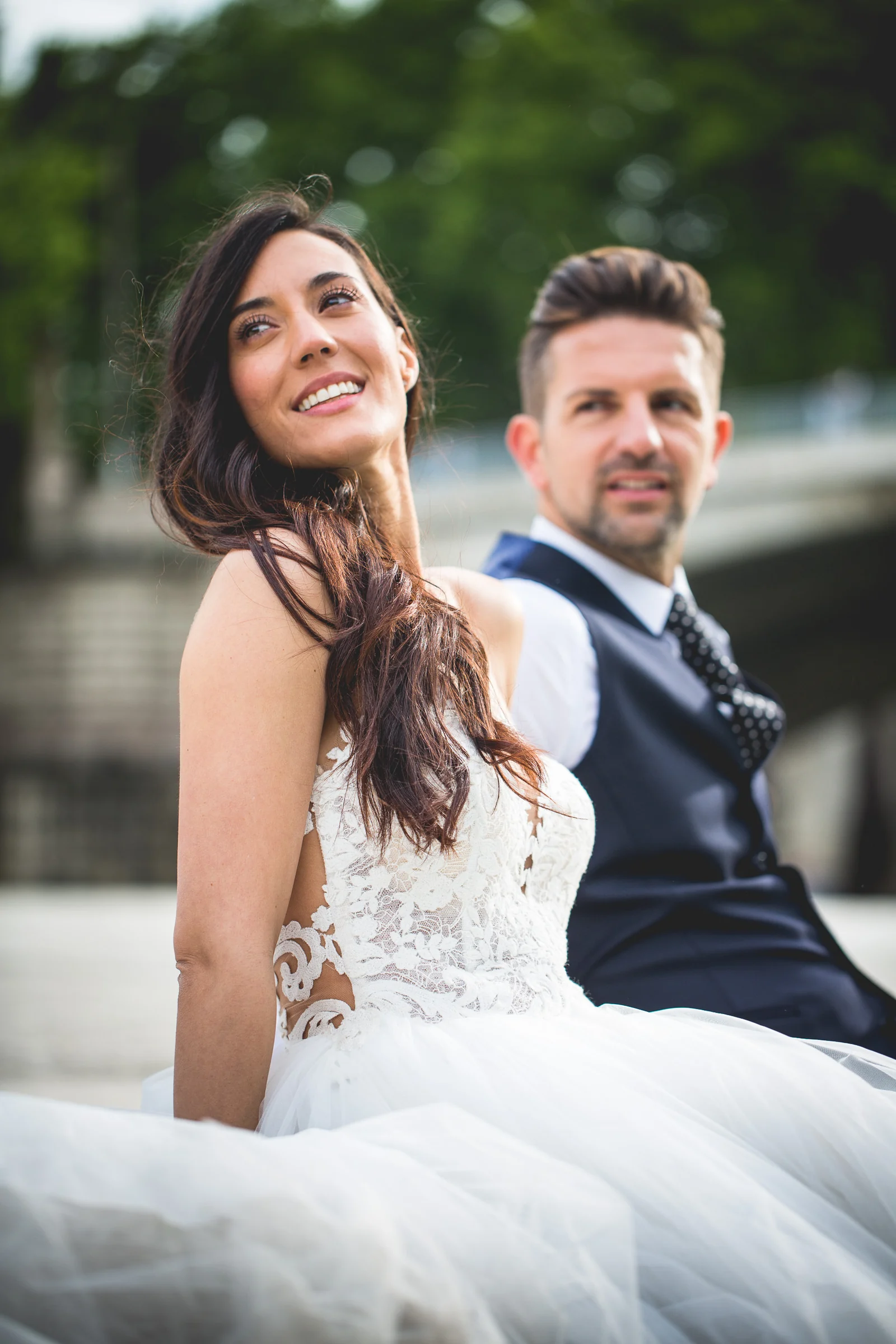 Retrato vertical de la pareja sentada y sonriente junto al Tíber durante una sesión postboda en Río Tíber y puentes de Roma.