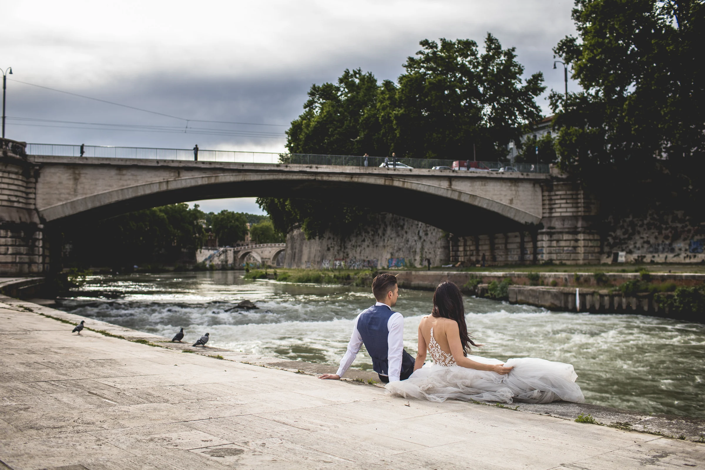 Los novios sentados frente al río con un puente al fondo durante una sesión postboda en Río Tíber y puentes de Roma.