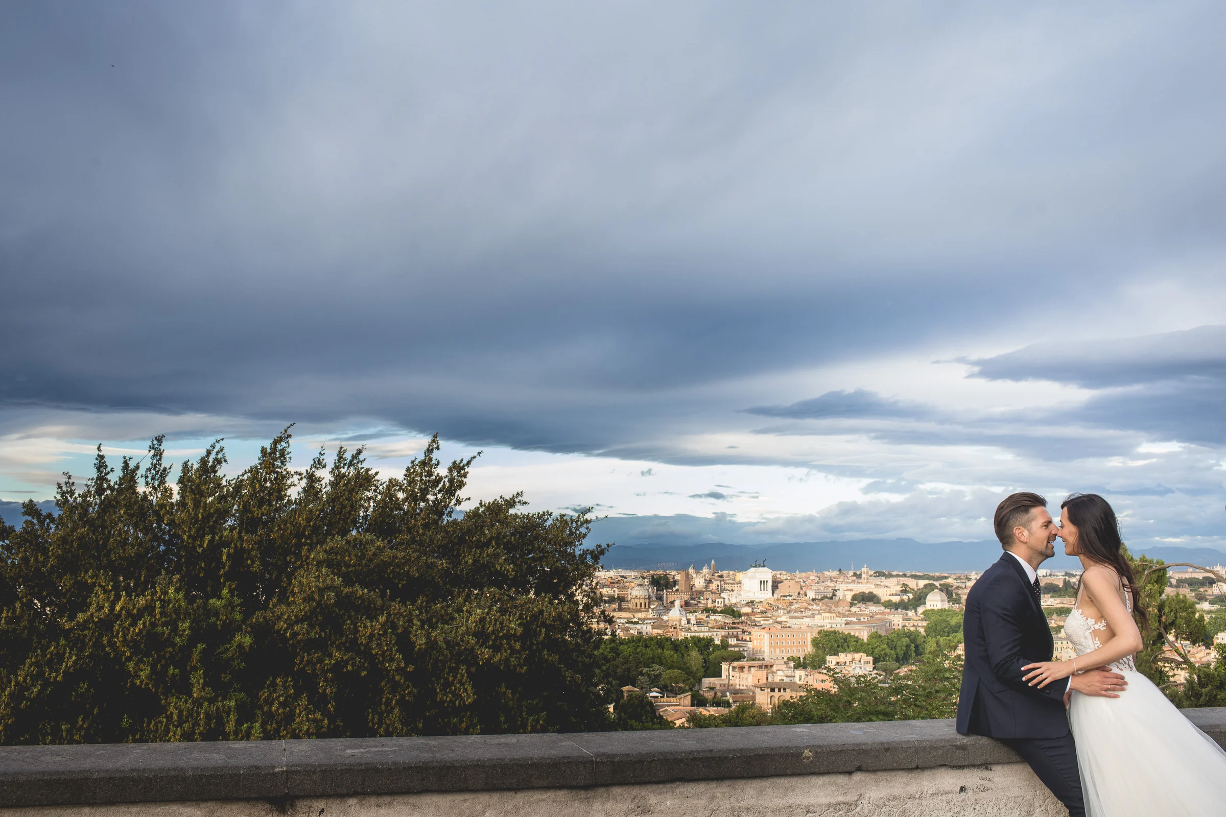Los novios se abrazan bajo un cielo dramático con la ciudad al fondo durante una sesión postboda en Miradores y centro histórico de Roma.