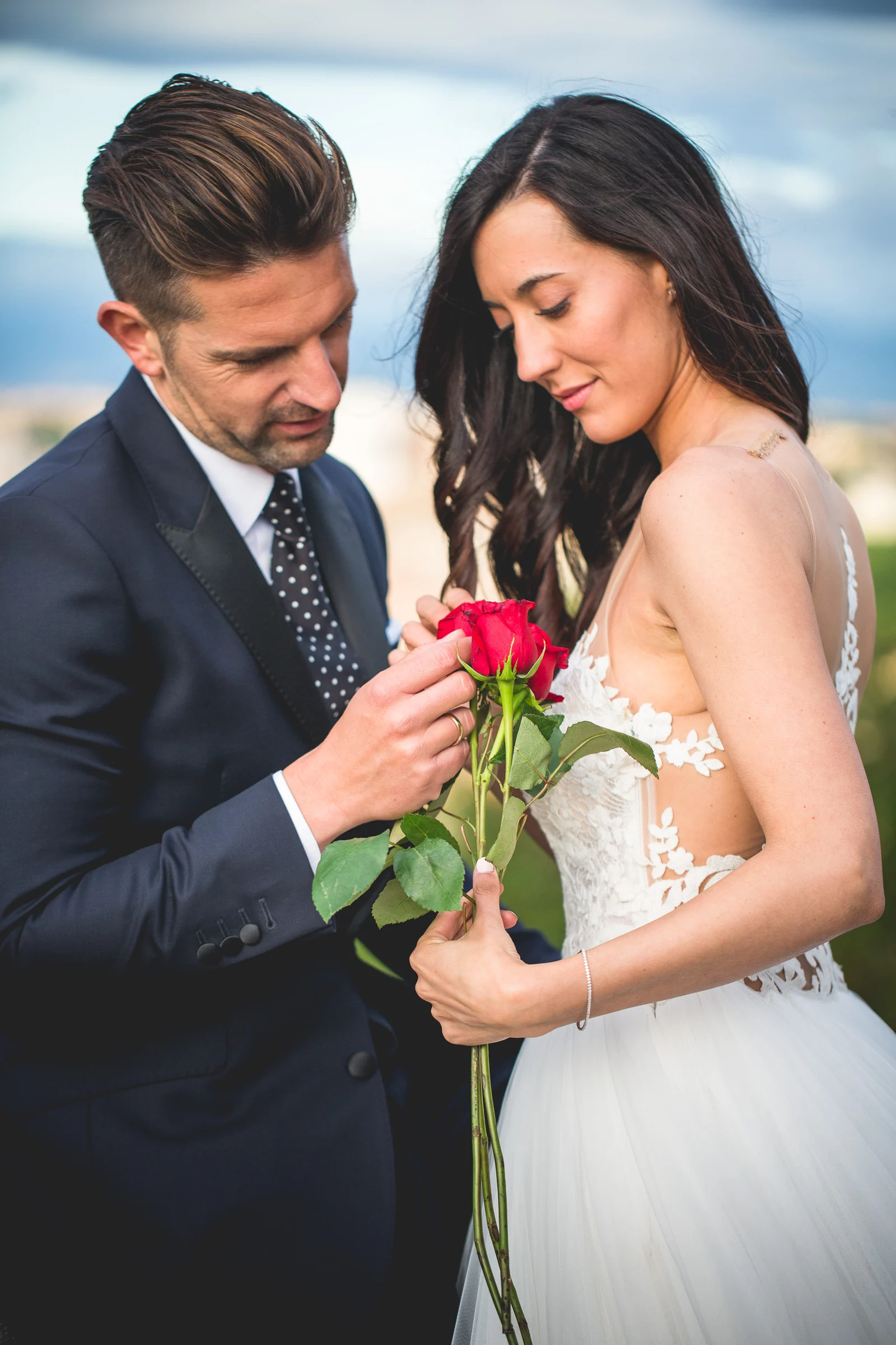 Los novios observan juntos una rosa roja en un retrato vertical durante una sesión postboda en Miradores y centro histórico de Roma.