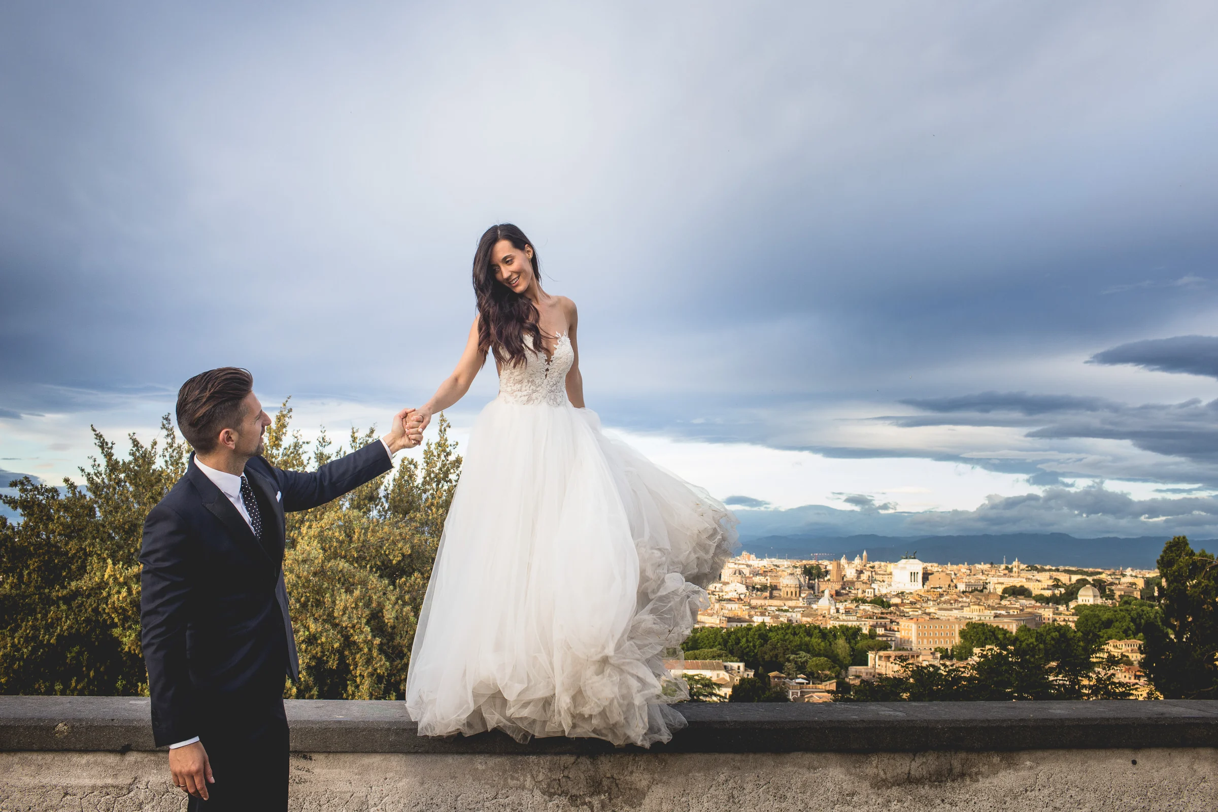 La novia posa elevada sobre un muro mientras el novio la mira desde abajo durante una sesión postboda en Miradores y centro histórico de Roma.