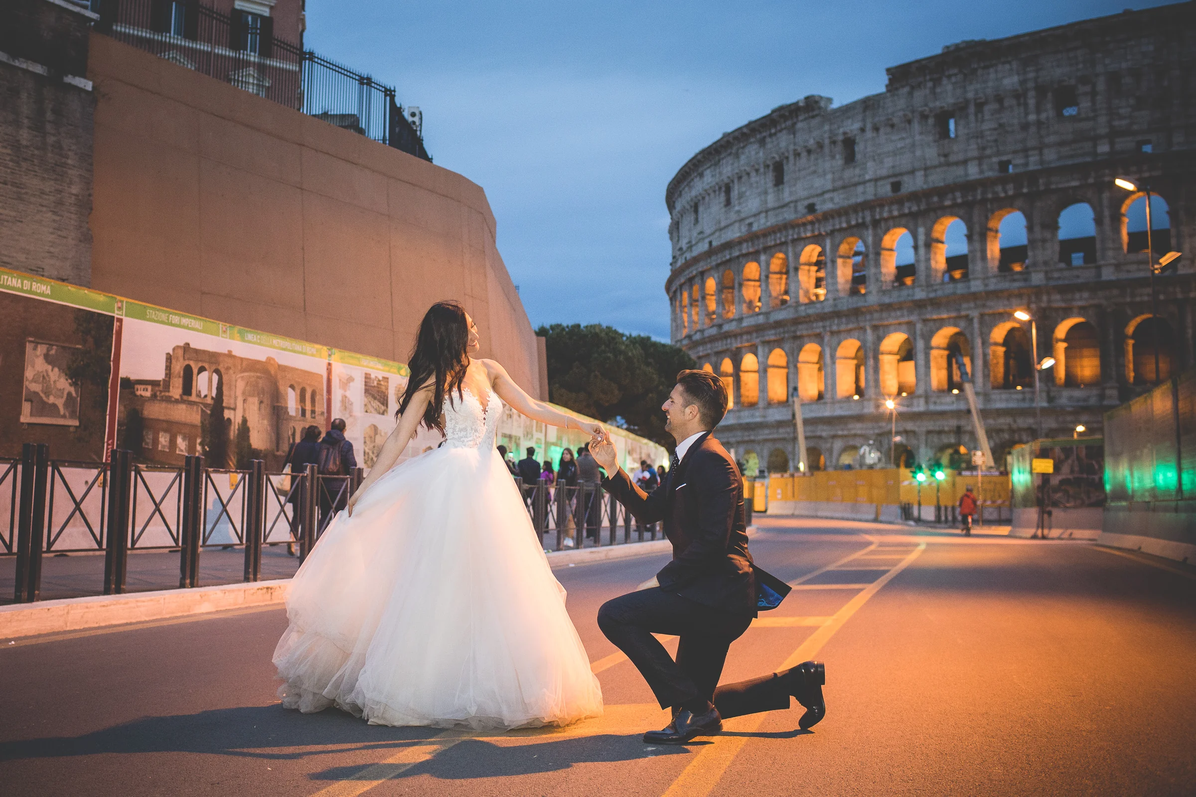 El novio se arrodilla ante la novia con el Coliseo iluminado al fondo durante una sesión postboda en Coliseo de Roma de noche.