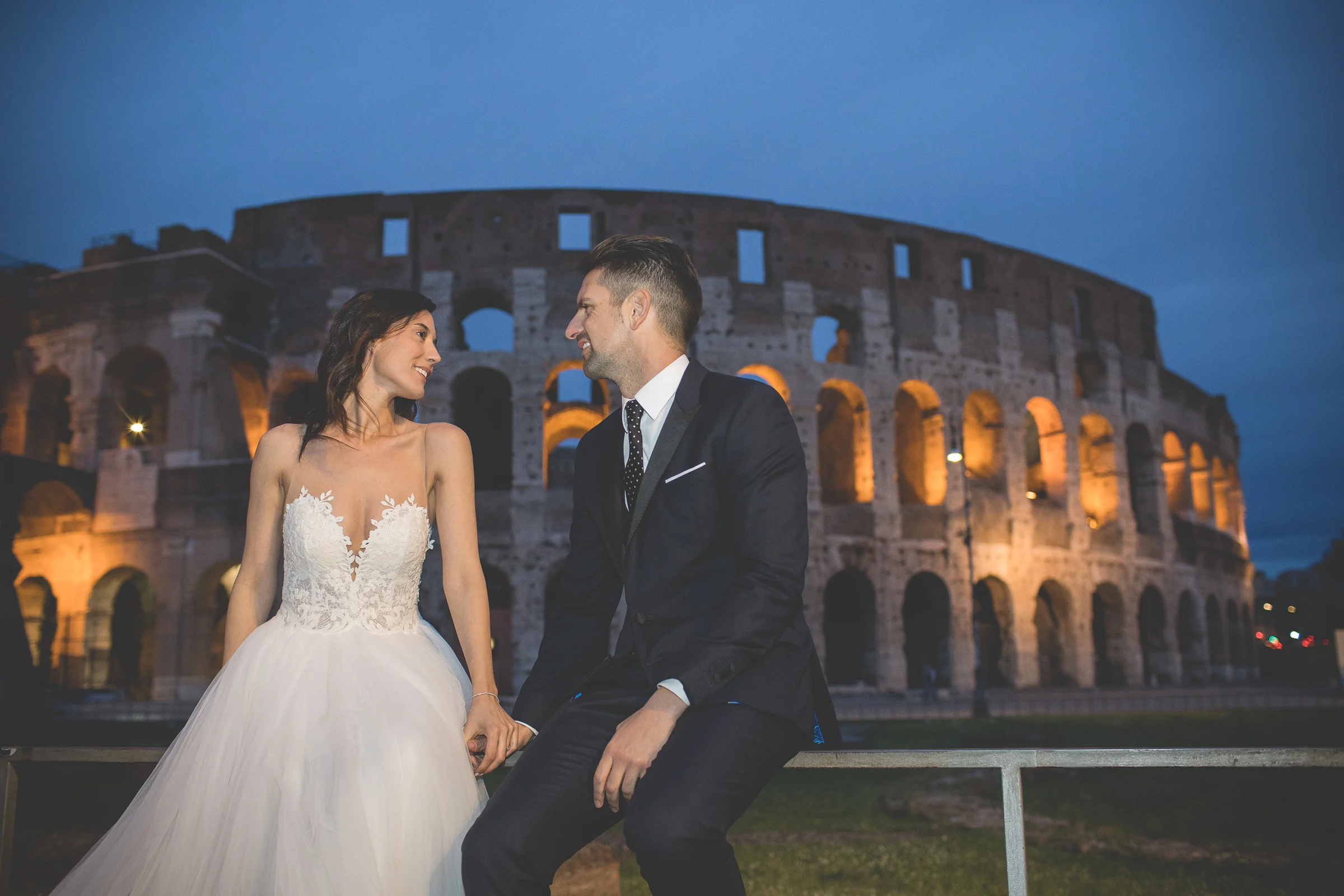 Los novios se miran de noche con el Coliseo iluminado detrás durante una sesión postboda en Coliseo de Roma de noche.