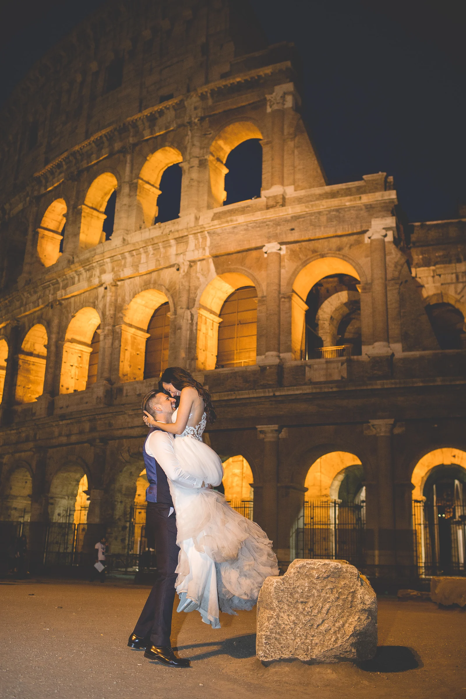 El novio levanta a la novia frente al Coliseo iluminado durante una sesión postboda en Coliseo de Roma de noche.