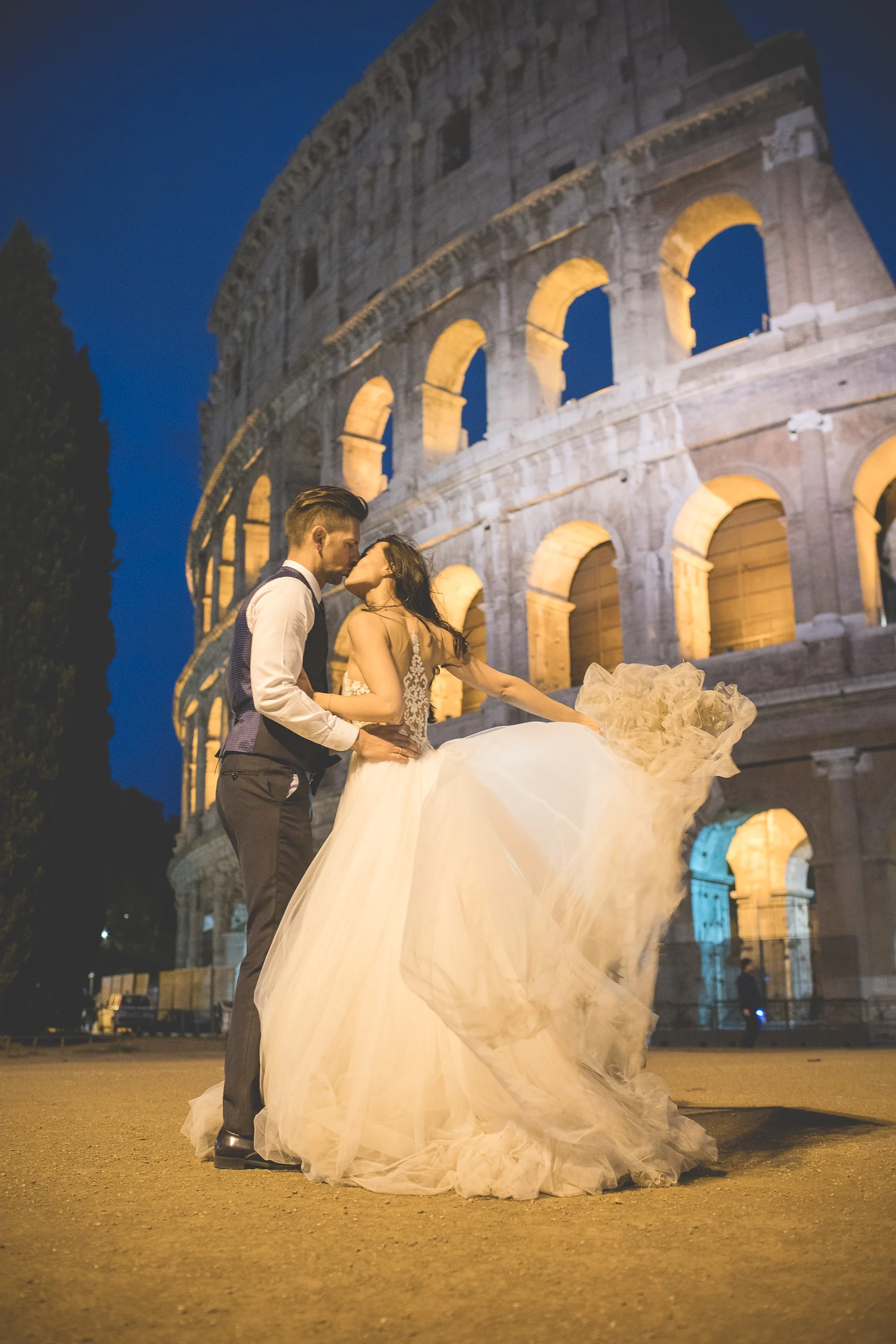 Los novios se besan con el vestido en movimiento ante el Coliseo nocturno durante una sesión postboda en Coliseo de Roma de noche.
