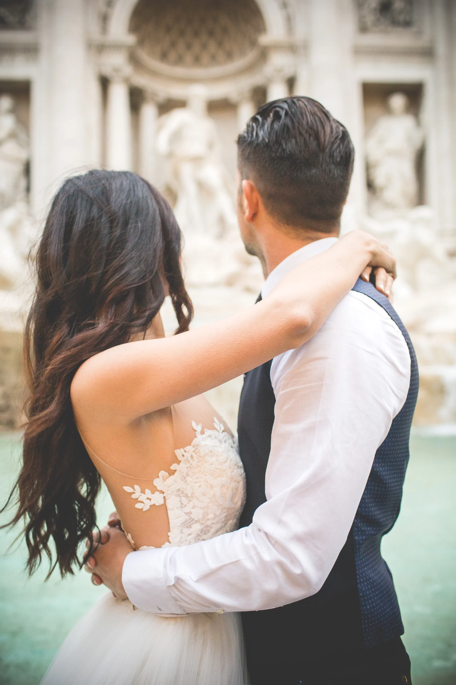 Los novios sentados de espaldas junto al agua de la Fontana di Trevi durante una sesión postboda en Fontana di Trevi.